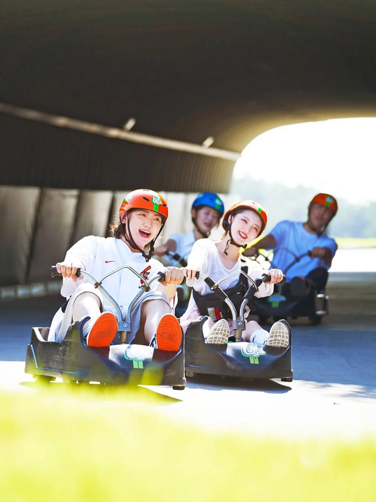 Four people wearing colorful safety helmets riding Skyline Luge carts through a tunnel in Busan, expressing joy and excitement.