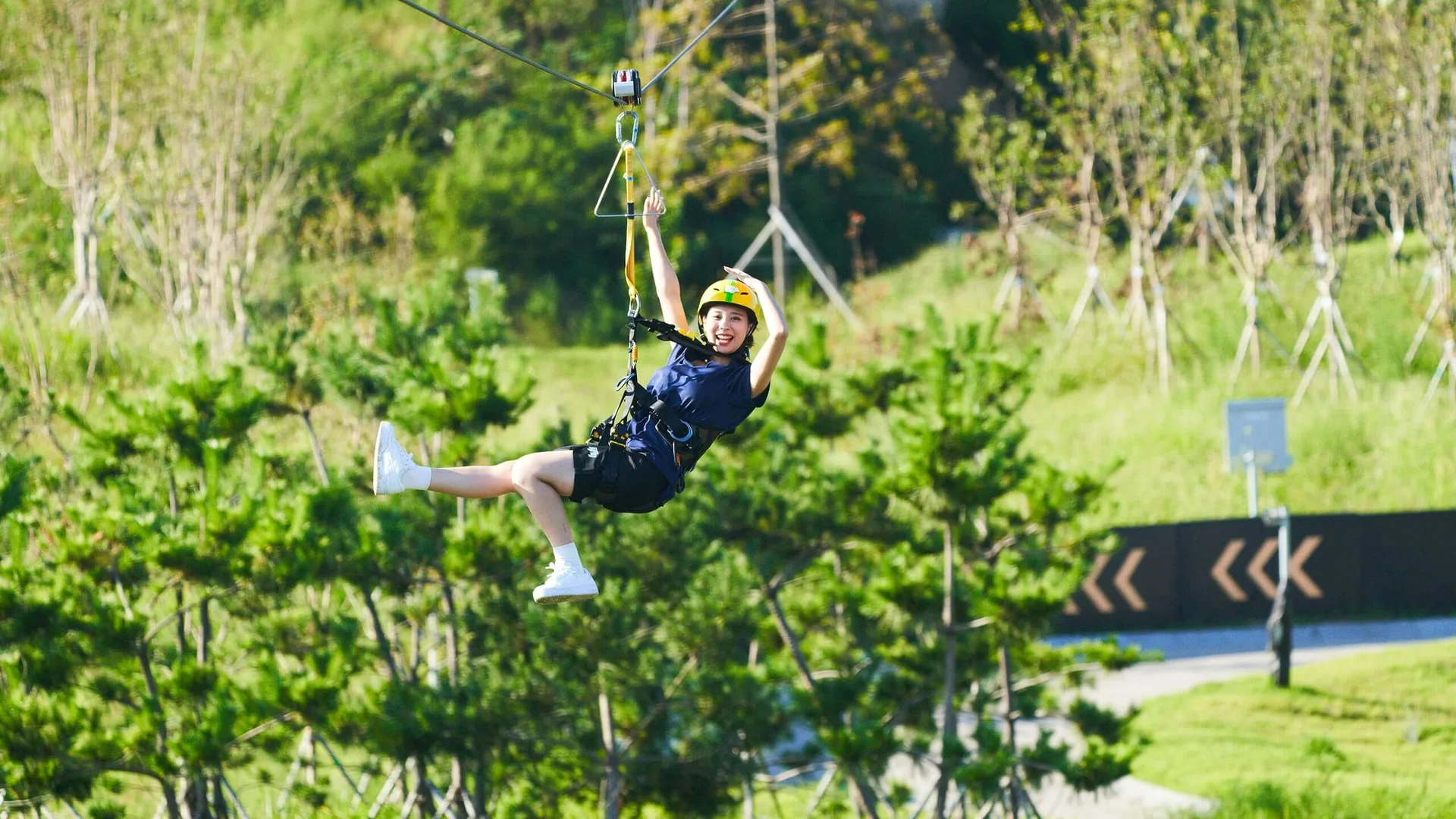 A woman wearing a yellow helmet and safety harness gliding on a zipline at Skyline Luge Busan, surrounded by green trees.