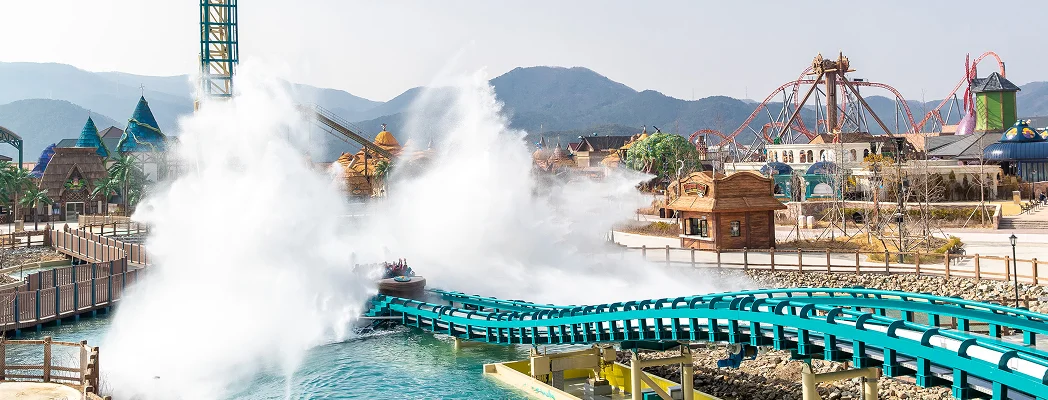 A wide-angle, eye-level shot of the Giant Splash attraction at Lotte World Adventure Busan. In the center, a ride vehicle has just plunged into a teal-colored track's splash zone, creating a massive, towering explosion of white water that erupts upward and outward. The track is supported by light-colored pillars over a turquoise lagoon.  In the background, the park's landscape features a mix of whimsical, storybook-style buildings with blue conical roofs and the skeletal orange structures of other thrill rides like the Giant Swing and Giant Digger roller coaster. The entire scene is set against a backdrop of hazy, rolling mountains under a bright, clear sky, capturing the scale and excitement of the theme park.