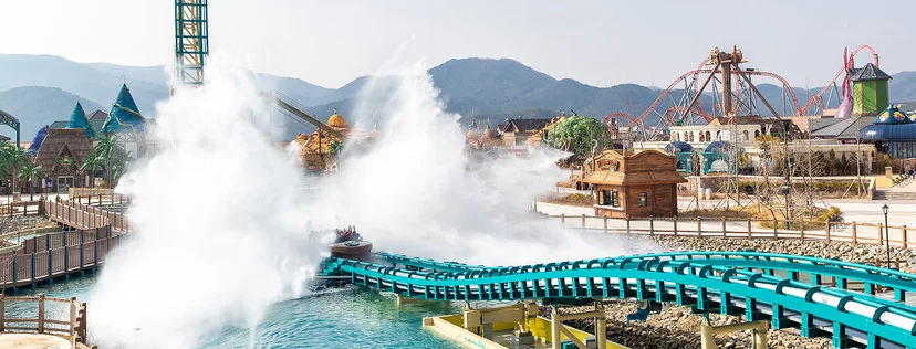 A wide-angle, eye-level shot of the Giant Splash attraction at Lotte World Adventure Busan. In the center, a ride vehicle has just plunged into a teal-colored track's splash zone, creating a massive, towering explosion of white water that erupts upward and outward. The track is supported by light-colored pillars over a turquoise lagoon.  In the background, the park's landscape features a mix of whimsical, storybook-style buildings with blue conical roofs and the skeletal orange structures of other thrill rides like the Giant Swing and Giant Digger roller coaster. The entire scene is set against a backdrop of hazy, rolling mountains under a bright, clear sky, capturing the scale and excitement of the theme park.