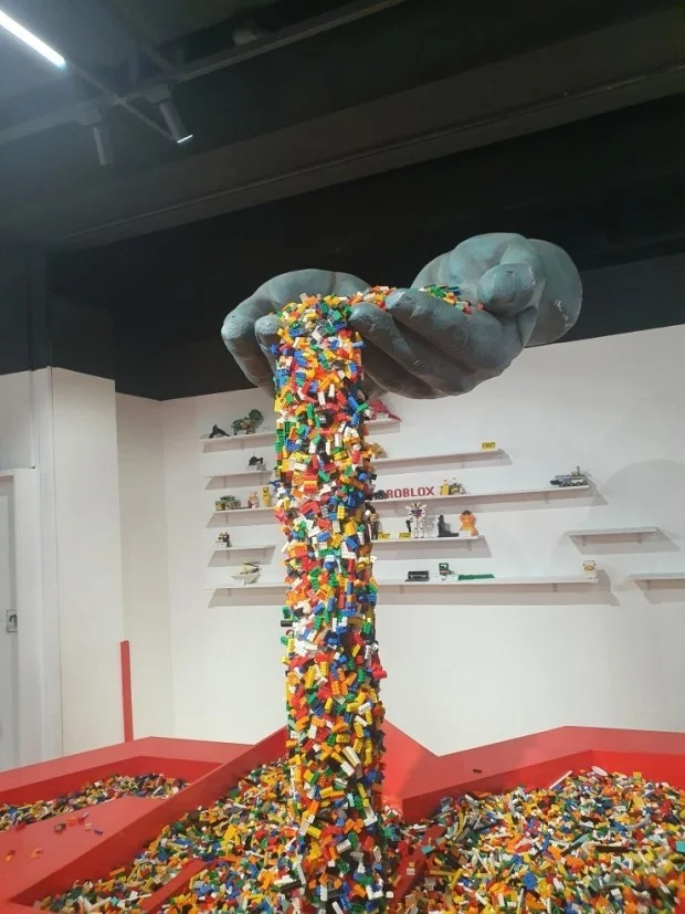  A vertical shot of a large-scale art installation at Brick Campus Busan. A pair of giant, gray sculpted hands emerges from a dark ceiling, cupped together as if pouring a waterfall of thousands of colorful toy bricks. The bricks—in vibrant shades of red, yellow, blue, green, and white—cascade downward in a thick, continuous stream into a large red pit filled with even more bricks.  In the background, minimalist white walls feature thin shelves displaying various small brick models, including a "ROBLOX" sign and small character figures. The floor area is dominated by the red-bordered brick pit, creating a playful and immersive atmosphere that showcases the creative possibilities of brick art.