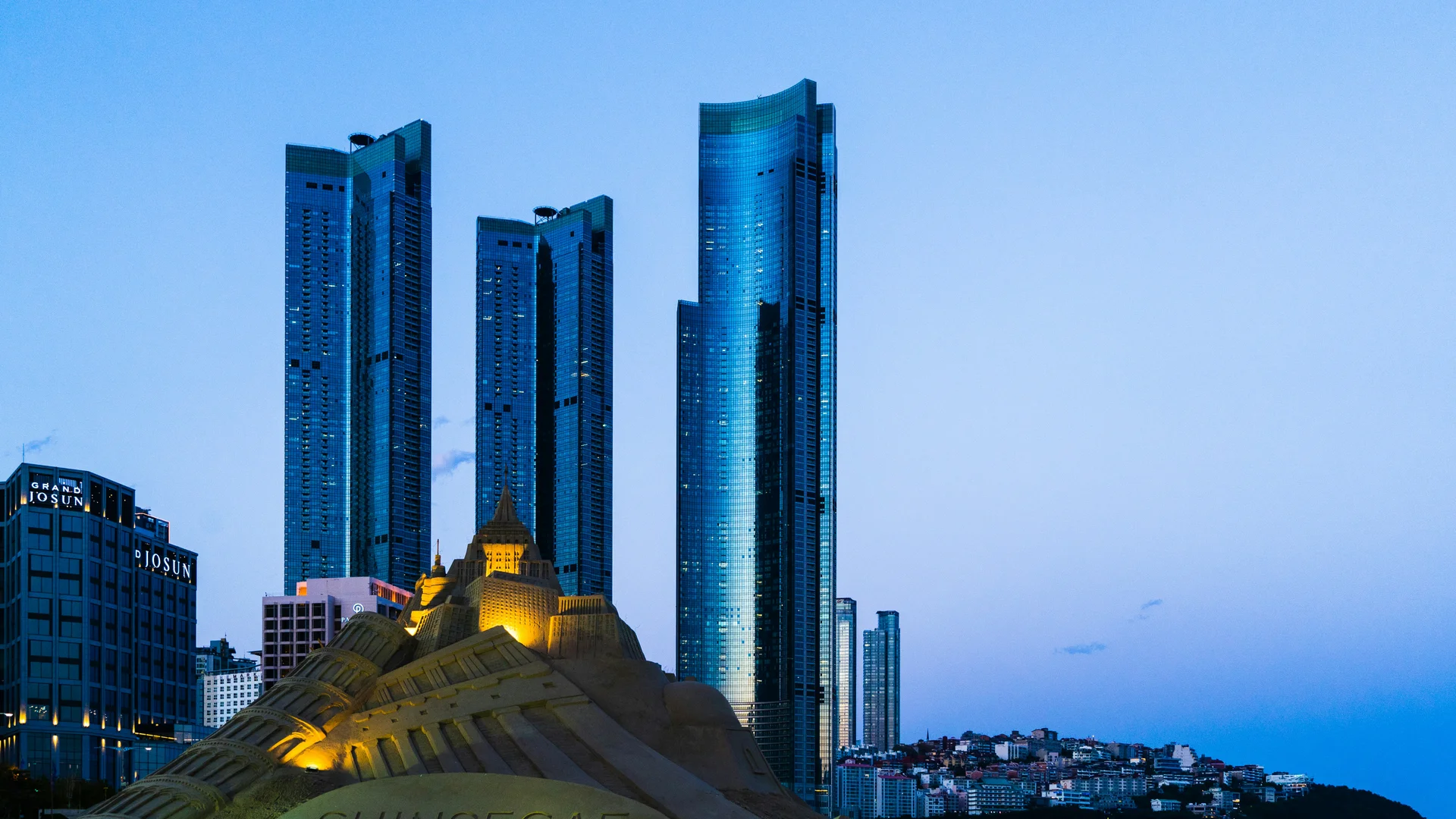A wide-angle landscape shot of Haeundae Beach in Busan at dusk. In the foreground, a large, intricately carved sandcastle sculpture is illuminated by warm spotlights, showing detailed architectural features like towers and windows.  Behind the sculpture, the skyline is dominated by three glowing, blue-tinted glass skyscrapers (Haeundae LCT The Sharp) that stretch toward a deep blue twilight sky. To the left, the "Grand Josun" hotel building is visible, and to the right, the city lights of a hillside neighborhood begin to twinkle. The overall mood is calm and sophisticated, contrasting the organic texture of the sand with the sleek, modern urban architecture.