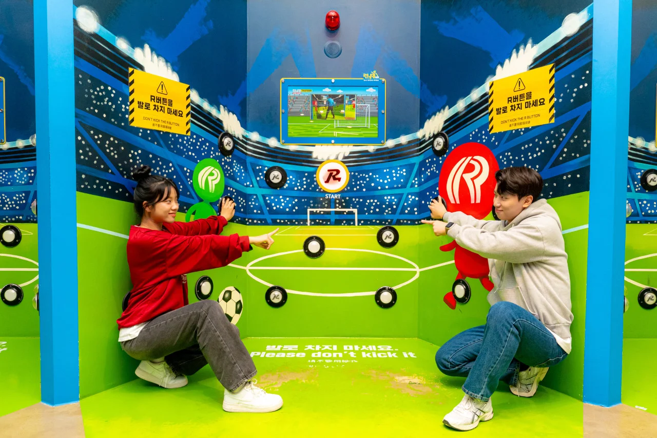 A vibrant, eye-level shot of an interactive game zone at Running Man Busan. Two people, a woman in a red sweatshirt and a man in a grey hoodie, are crouched on a bright green floor designed to look like a soccer field. They are both pointing playfully toward a central "START" button featuring the iconic Running Man "R" logo.  The walls are decorated with a colorful stadium-themed mural, complete with illustrated crowds and soccer goals. Several black circular touch-pads are scattered across the walls as part of the game. Two yellow warning signs in Korean and English advise players, "Please don't kick it." In the center, a digital screen displays a soccer-themed mini-game, and stylized red and green "R" character figures flank the players, adding to the energetic and immersive atmosphere of the attraction.
