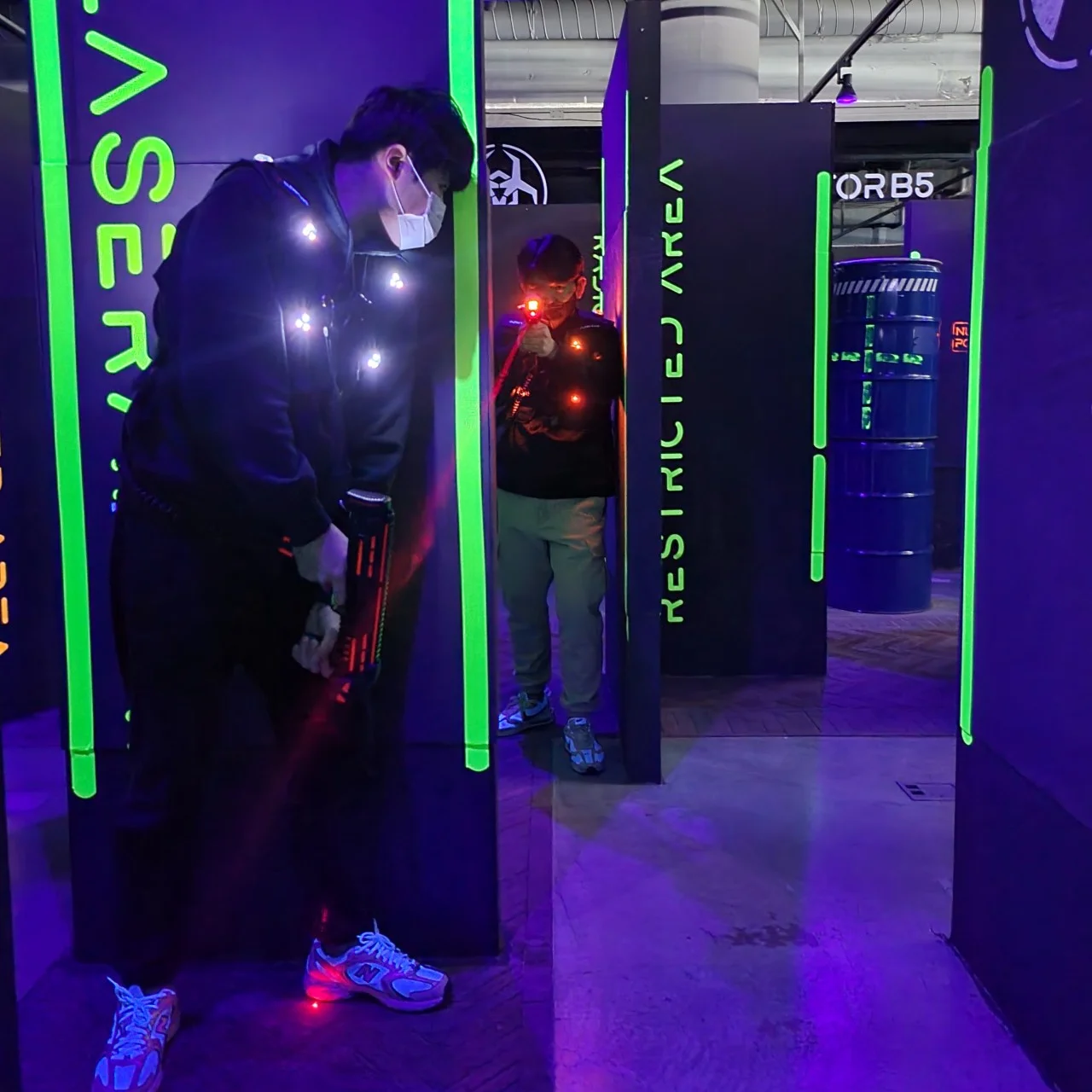 A medium-shot of two people engaged in a laser tag game at Laser Arena Busan Samjeong. The setting is a dark, indoor arena illuminated by vibrant neon green lights on black partition walls labeled with text like "RESTRICTED AREA."  In the foreground, a person wearing a black hoodie, a face mask, and glowing sensors on their vest stands in a defensive posture, holding a laser gun. In the background, another player is partially visible behind a partition, aiming a laser gun that emits a bright red beam toward the foreground. The floor has a purple hue from the ambient neon lighting, creating an intense, high-energy atmosphere for an indoor action activity.