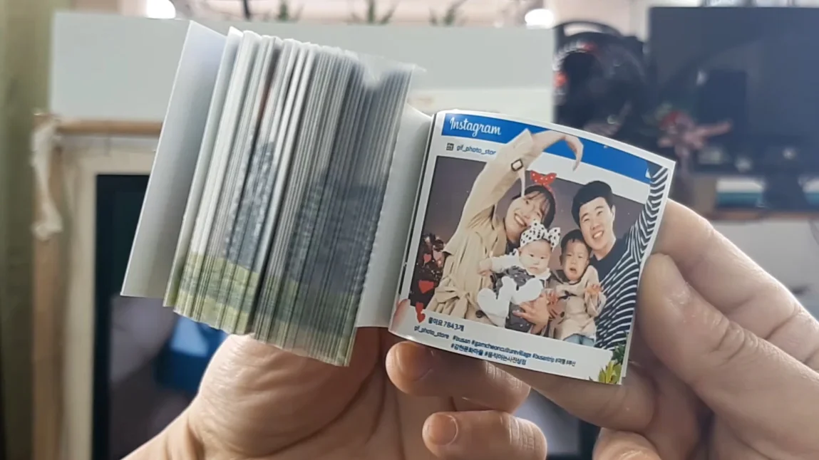 A close-up shot of a pair of hands holding an open flipbook from the Moving Photo Store (움직이는 사진상점) in Gamcheon Culture Village. The flipbook is designed to look like an Instagram post, featuring a photo of a smiling family—a mother, father, and two young children—against a night sky backdrop.  The left side of the book shows the thick edge of numerous pages that, when flipped, create a moving animation of the family's poses. The right side displays the clear, final frame of the "post," complete with the Instagram logo, a username, and hashtags like #busan and #gamcheonculturevillage at the bottom. The background is slightly blurred, focusing all attention on the personalized souvenir.