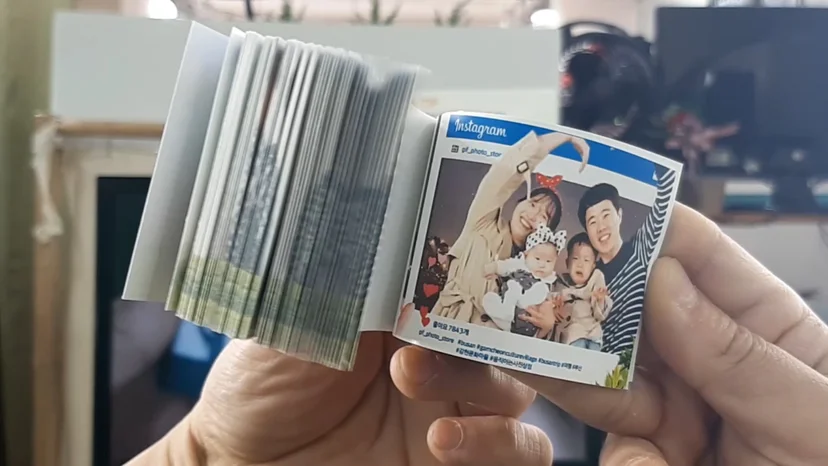 A close-up shot of a pair of hands holding an open flipbook from the Moving Photo Store (움직이는 사진상점) in Gamcheon Culture Village. The flipbook is designed to look like an Instagram post, featuring a photo of a smiling family—a mother, father, and two young children—against a night sky backdrop.  The left side of the book shows the thick edge of numerous pages that, when flipped, create a moving animation of the family's poses. The right side displays the clear, final frame of the "post," complete with the Instagram logo, a username, and hashtags like #busan and #gamcheonculturevillage at the bottom. The background is slightly blurred, focusing all attention on the personalized souvenir.