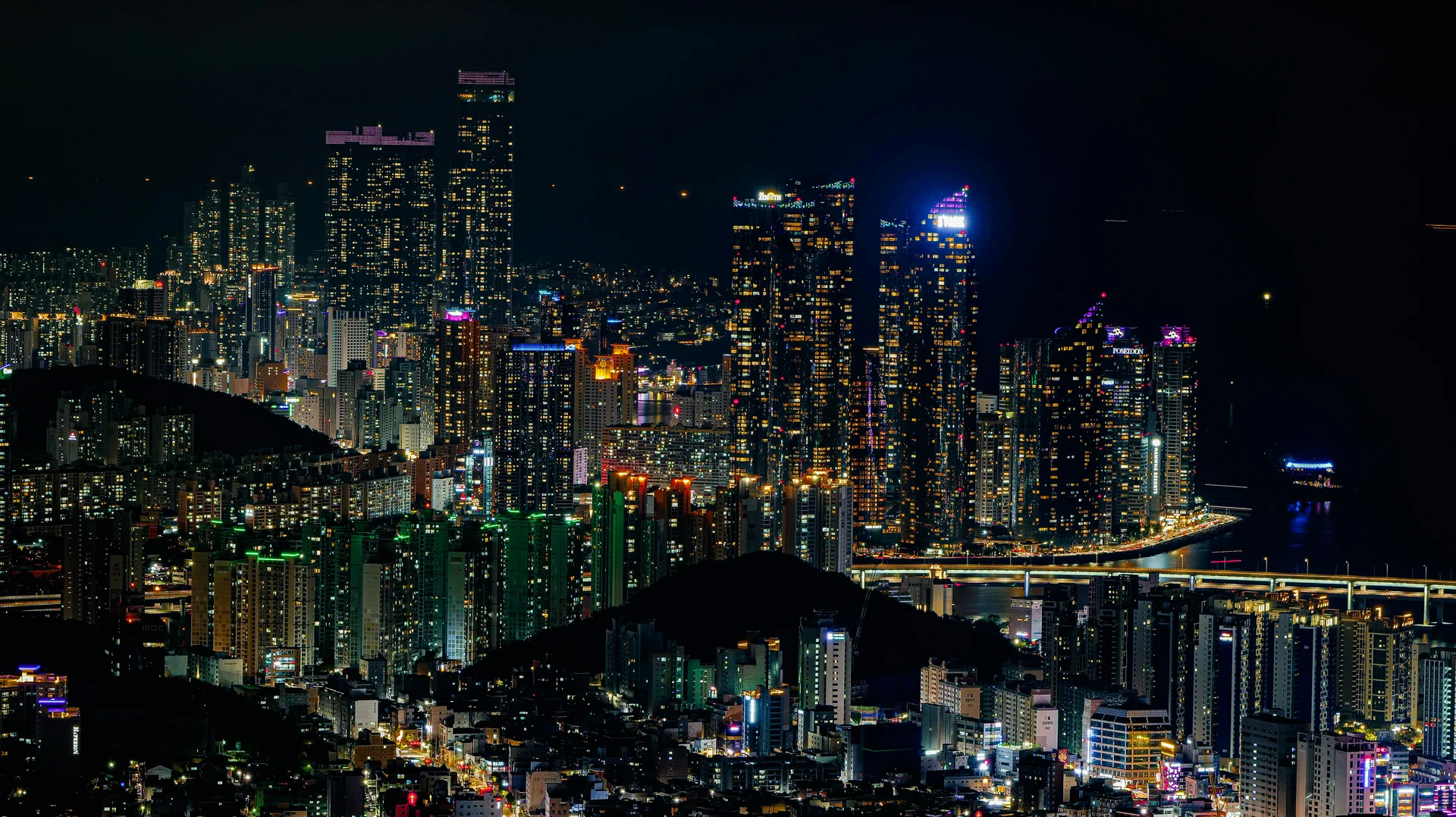  A wide-angle, low-angle landscape shot of the Marine City skyline in Haeundae, Busan, at night. The foreground features a calm, dark sea that perfectly reflects the vibrant, glowing lights of the surrounding architecture.  The skyline is dominated by several luxury high-rise skyscrapers, including the iconic curved glass towers of Haeundae I'Park and the towering Doosan We've the Zenith. The buildings are brilliantly illuminated with thousands of white and blue internal lights, creating a "festival of light" that mimics the aesthetic of major global cities like Hong Kong or Shanghai.  On the right, the Gwangan Bridge (Diamond Bridge) stretches across the water, its double-deck suspension structure glowing with bright LED lights. The sky is a deep, clear indigo, providing a sharp contrast to the shimmering gold and blue reflections dancing on the water's surface. The overall atmosphere is modern, sophisticated, and energetic, capturing Busan's status as a premier global port city.