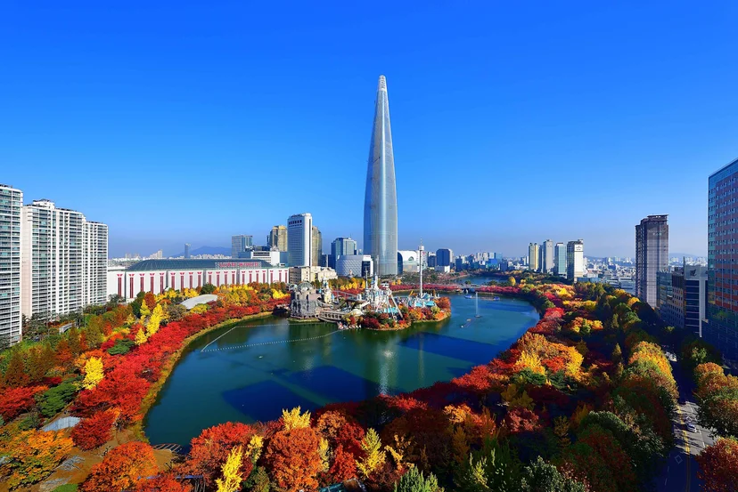 Aerial view of Seokchon Lake autumn foliage in Seoul, featuring vibrant red and orange leaves surrounding Lotte World Magic Island and the towering Lotte World Tower under a clear blue sky.