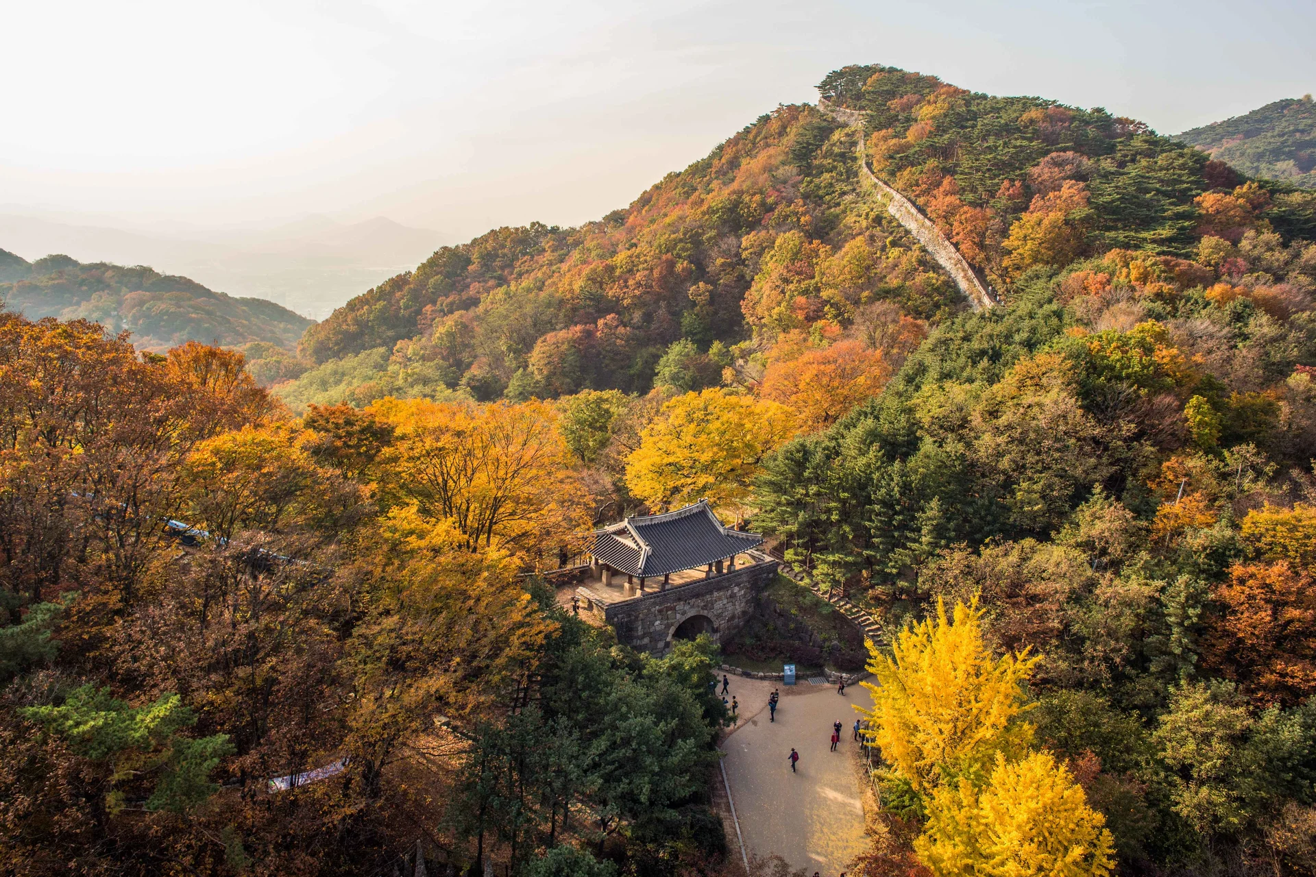 Scenic autumn view of Namhansanseong Fortress, a UNESCO World Heritage site in Gyeonggi-do, South Korea. The image features vibrant orange and yellow fall foliage surrounding the historic stone walls and gate, with hikers walking along the trail near Seoul.