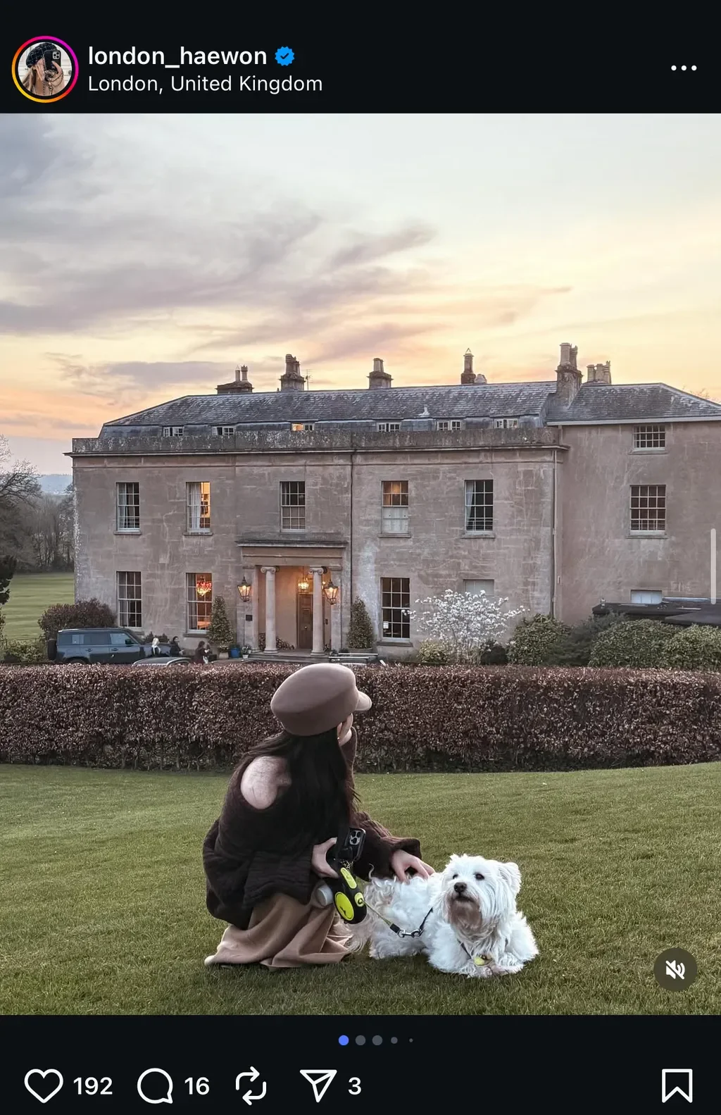 노을 지는 분홍빛 하늘 아래, 웅장한 고풍스러운 저택 앞 잔디밭에서 갈색 베레모와 스웨터를 입은 여성이 하얀 강아지와 함께 여유로운 시간을 보내고 있는 모습의 인스타그램 게시글