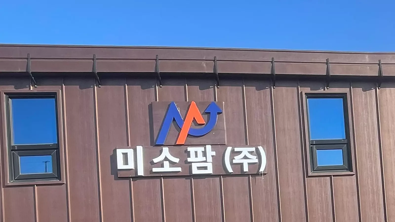 미소팜 주식회사 건물 외벽에 회사 로고와 이름이 크게 부착되어 있고, 청명한 하늘 아래 두 개의 창문이 위치한 모습