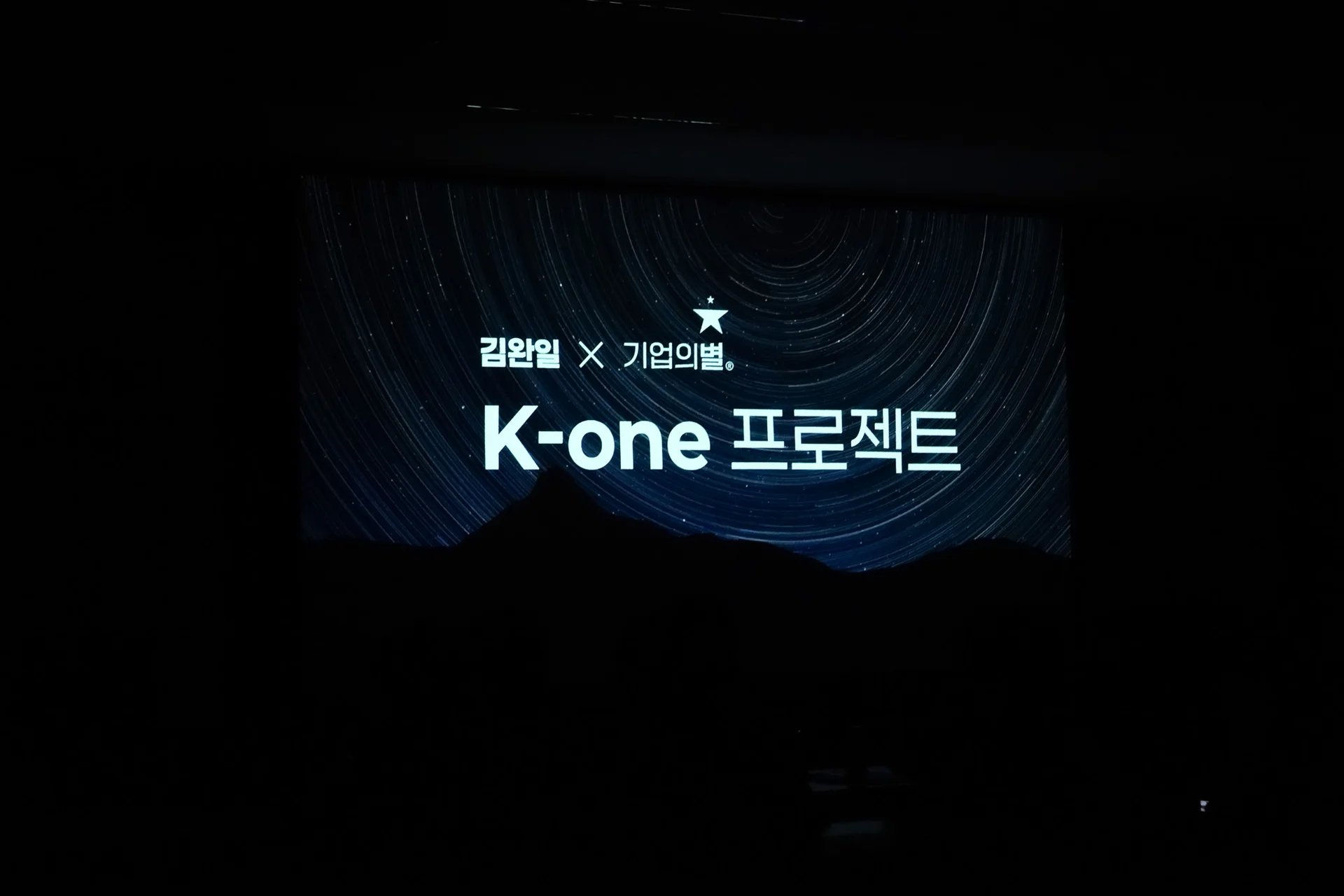 인트로 영상 사진 1. 암전된 실내에서 스크린에 김완일x기업의별 K-one 프로젝트라는 텍스트 로고가 크게 떠있으며 배경으로 별 궤적과 산이 있는 이미지가 띄워져있다.