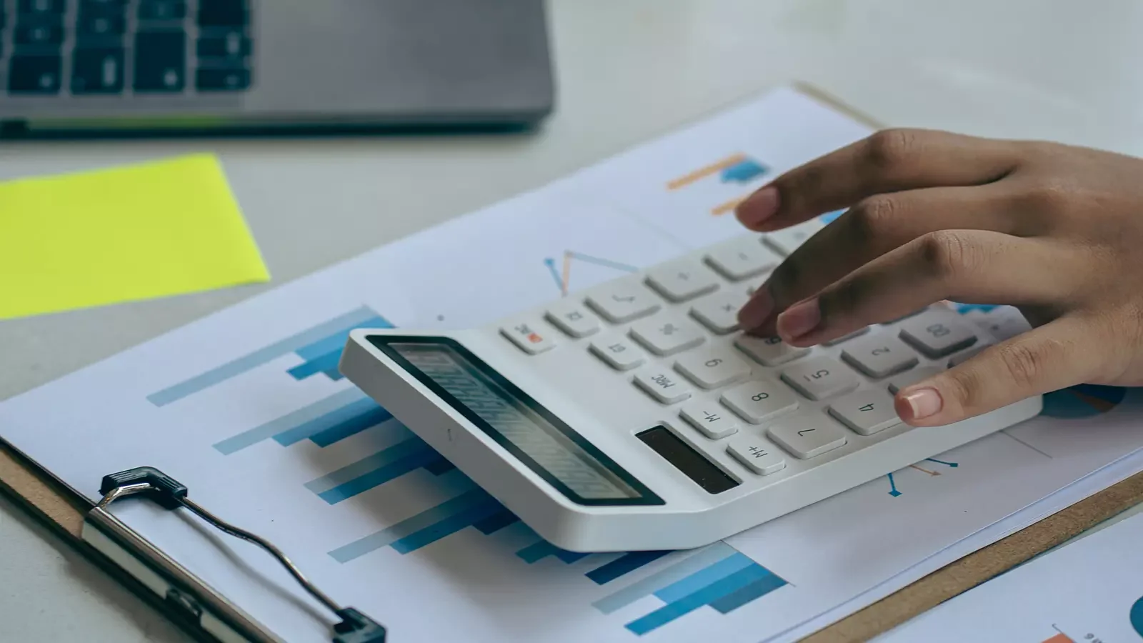 재무 자료와 그래프가 인쇄된 문서 위에서 계산기를 사용하는 손의 모습으로, 제조업 비과세 관련 비용 산정과 회계 처리 업무가 이루어지는 상황