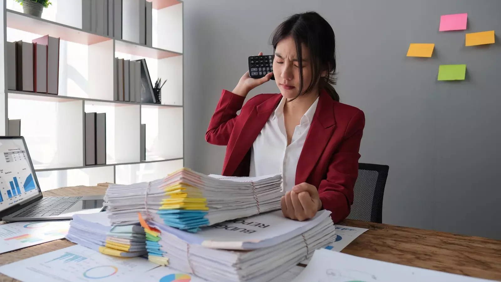 서류 더미와 노트북이 놓인 책상에서 계산기를 사용하며 비품 회계처리를 진행 중인 사무실 직원의 모습