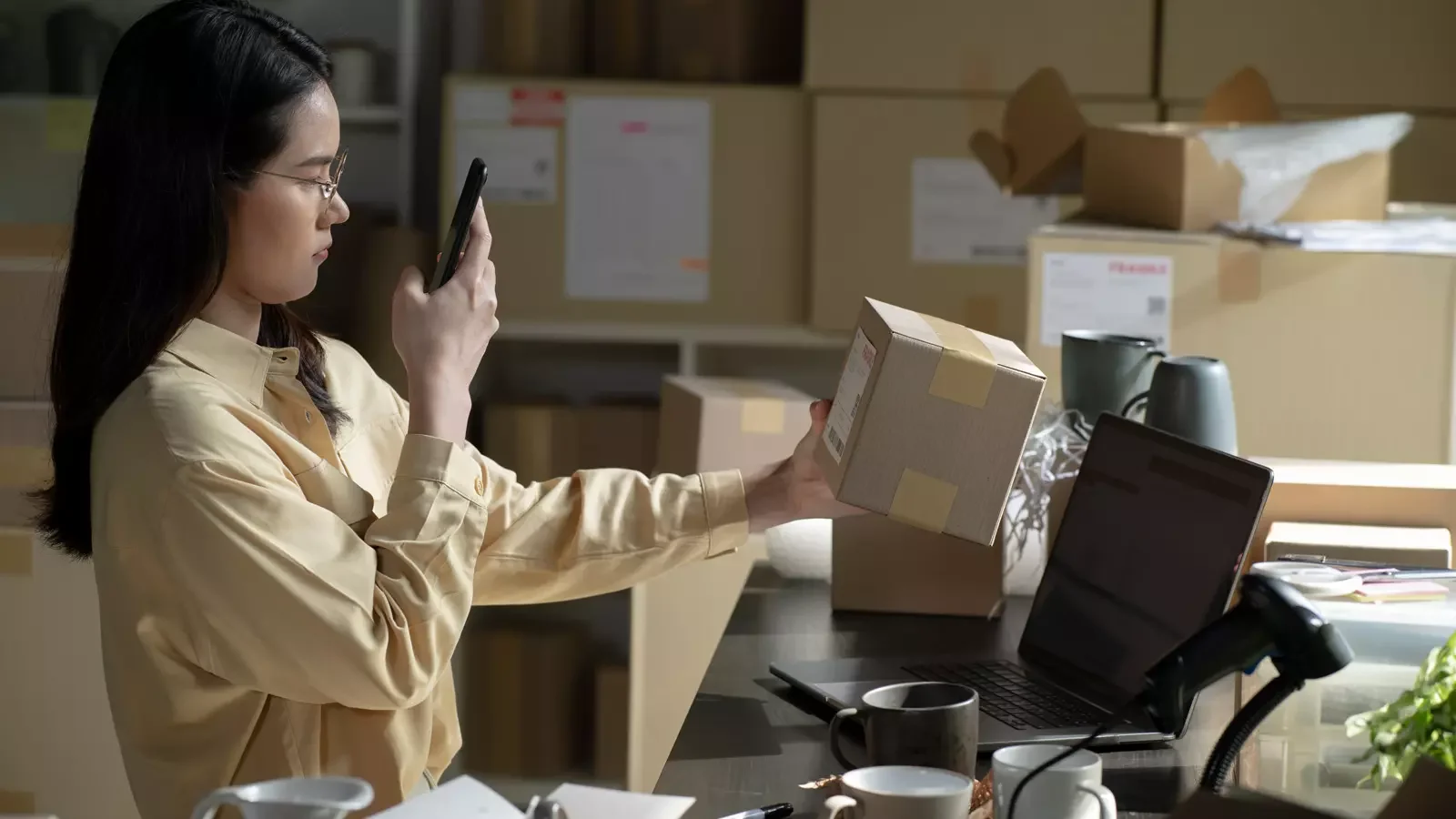 사무실 환경에서 여성이 스마트폰 카메라로 택배 박스를 스캔하며 재고를 검수하는 모습. 모바일 기기와 노트북을 연동한 스마트 물류 및 이커머스 재고 관리 프로세스