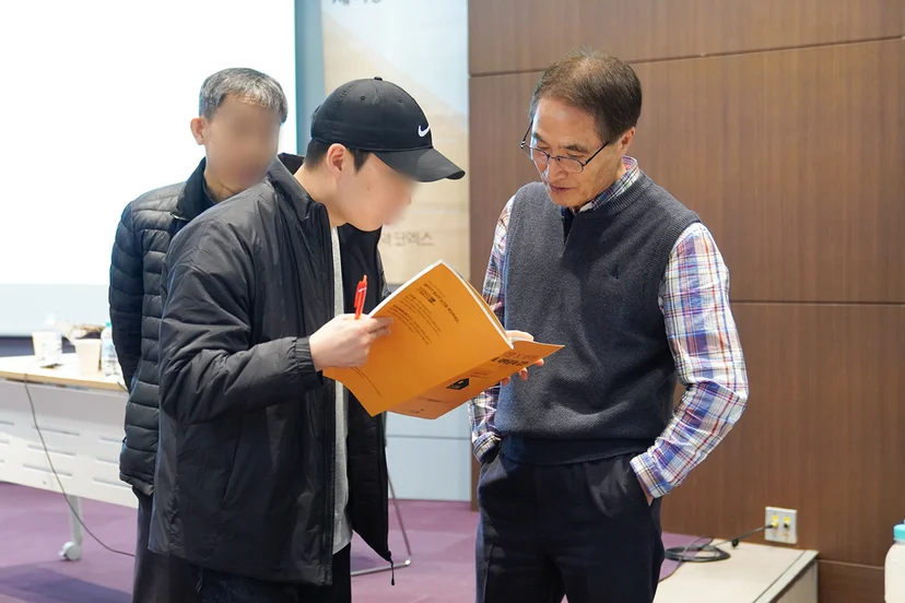 캡모자를 쓴 젊은 남성 세무사가 교재를 들고 배택현 세무사에게 질문하는 사진