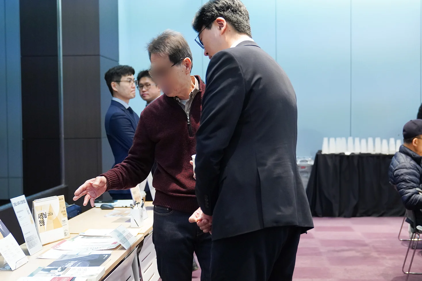 와인색 겉옷을 입은 중년의 남성 세무사가 부스를 손으로 가리키고 기업의별 스탭이 설명하는 모습