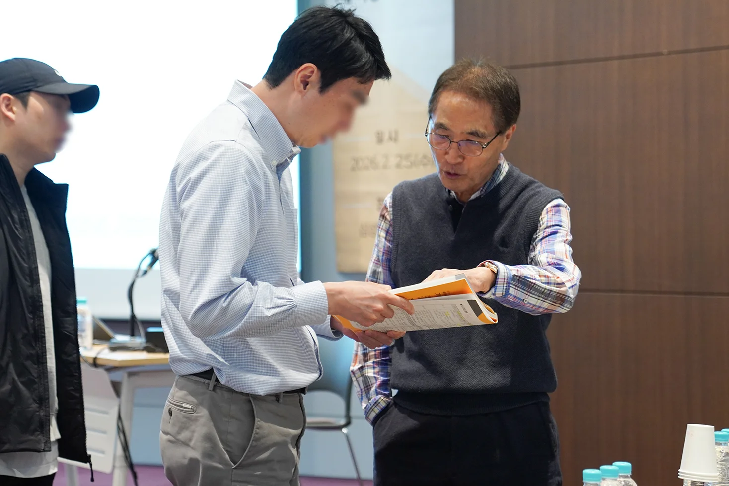 청색 작은 격자무늬 셔츠를 입은 젊은 남성 세무사가 교재를 들고, 배택현 세무사가 교재를 손으로 짚으며 설명하는 사진