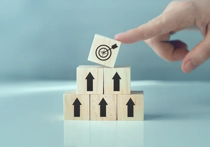 목표 아이콘이 그려진 나무 블록을 위로 쌓아 올리며 단계적으로 목표 달성을 상징하는 이미지