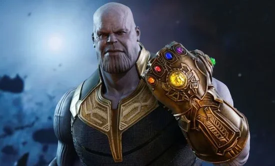 A powerful purple-skinned villain stands in a dramatic cosmic setting, wearing dark blue and gold armor and raising a large golden gauntlet embedded with six glowing multicolored stones. The character has a bald head, a stern expression, and a massive muscular build. The background is dark and space-like, with a soft blue light creating a cinematic atmosphere. This image evokes themes of superhero movies, fantasy villains, cosmic power, and epic battle scenes. Keywords: supervillain, infinity gauntlet, superhero film, cosmic villain, fantasy action character.