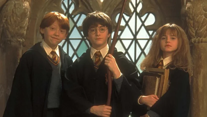 Portrait of three young students standing side by side in a stone hallway with large leaded windows behind them. They wear black robes over school uniforms with ties. One student holds a broomstick, another carries a stack of books, and all three face the camera with calm, confident expressions. The warm lighting and medieval-style architecture create a magical fantasy mood.