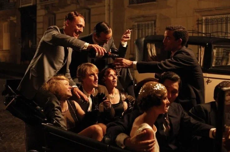 Scene from Midnight in Paris showing a lively group of partygoers in formal 1920s-style attire gathered in and around a car on a warmly lit Paris street at night.