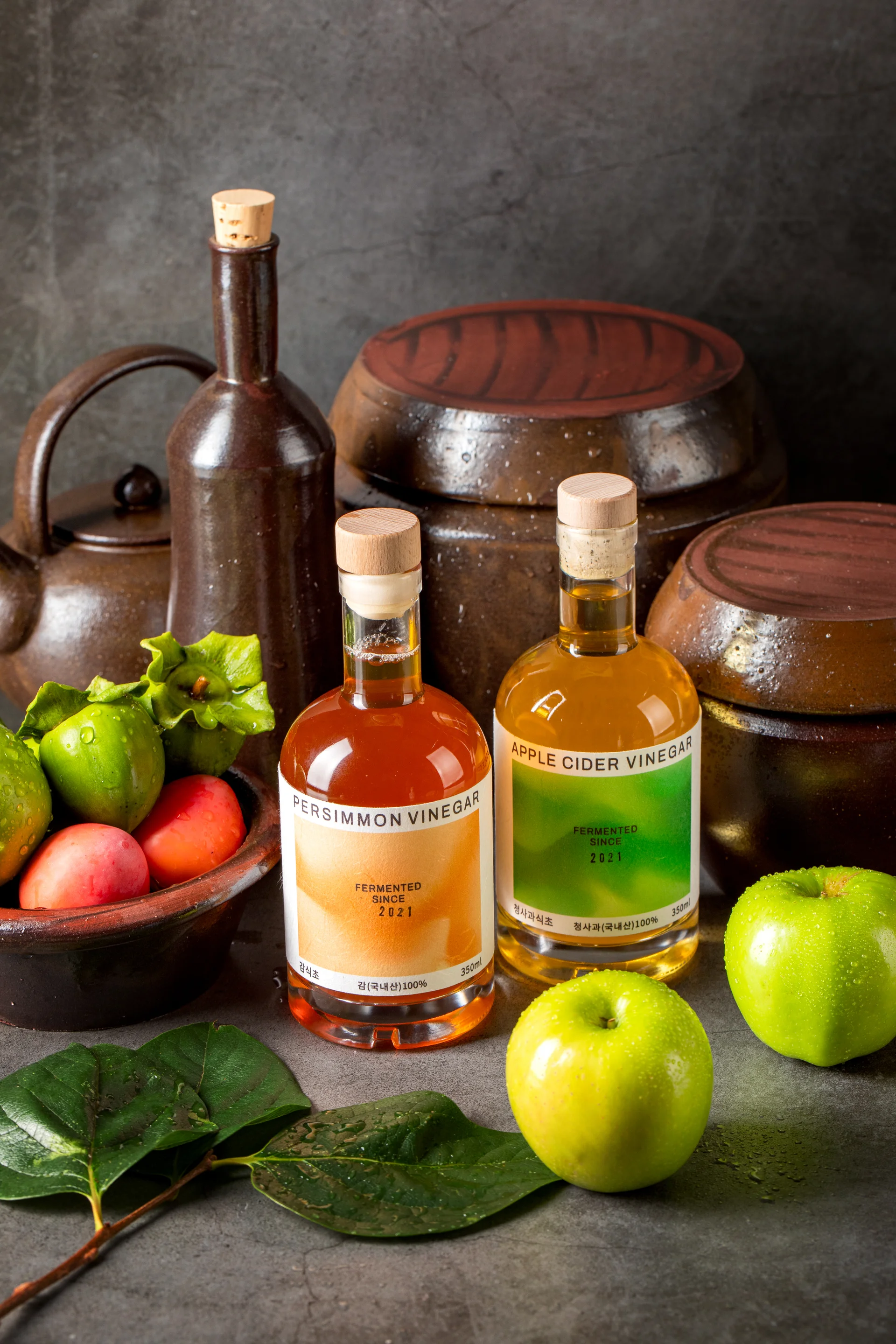 은가비 브랜드의 발효식초 3종(사과식초, 감식초, 식초 음료)이 신선한 사과와 감 원물과 함께 연출된 고감도 촬영 이미지. 한여름 감 수급 변수를 극복하고 기획자가 직접 디렉팅한 내추럴 톤의 브랜드 비주얼 컷.