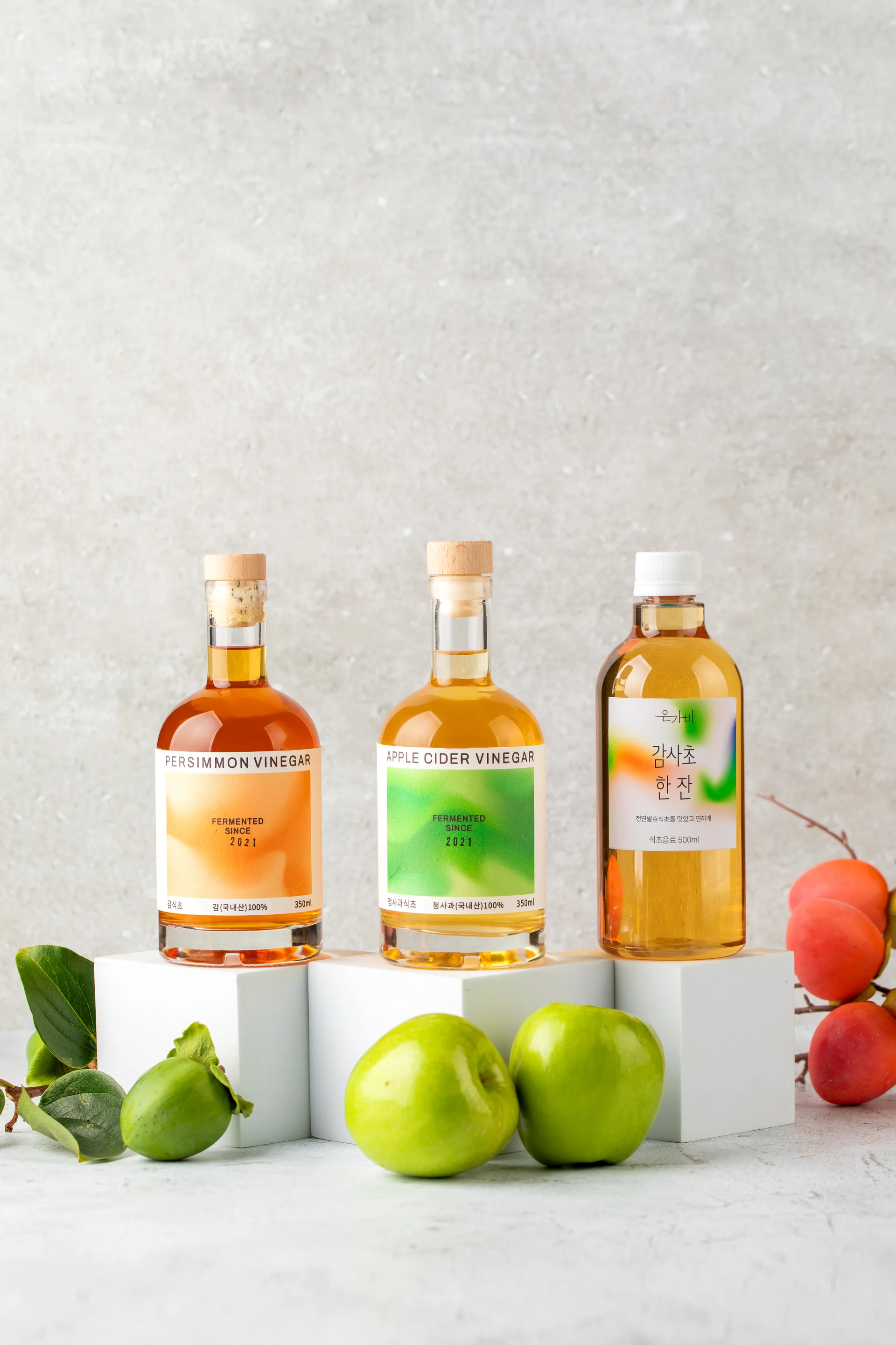 은가비 브랜드의 발효식초 3종(사과식초, 감식초, 식초 음료)이 신선한 사과와 감 원물과 함께 연출된 고감도 촬영 이미지. 한여름 감 수급 변수를 극복하고 기획자가 직접 디렉팅한 내추럴 톤의 브랜드 비주얼 컷.
