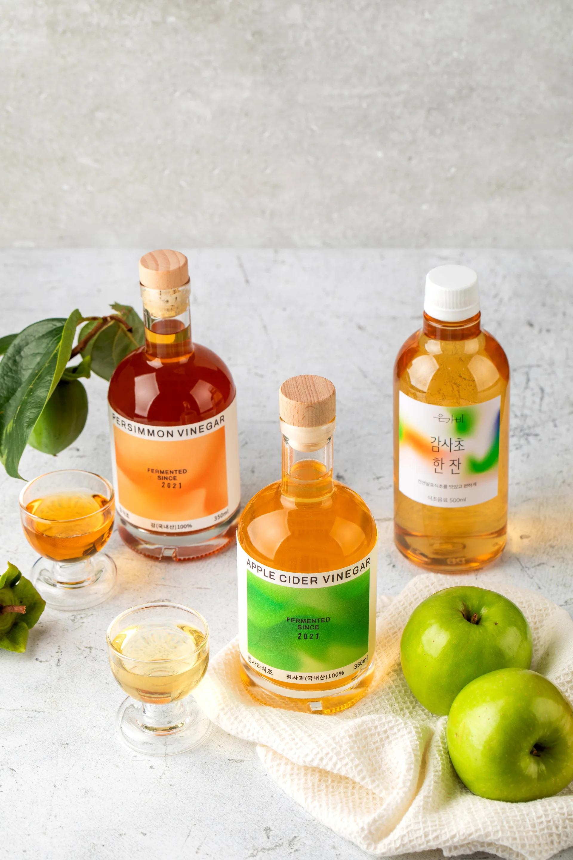은가비 브랜드의 발효식초 3종(사과식초, 감식초, 식초 음료)이 신선한 사과와 감 원물과 함께 연출된 고감도 촬영 이미지. 한여름 감 수급 변수를 극복하고 기획자가 직접 디렉팅한 내추럴 톤의 브랜드 비주얼 컷.