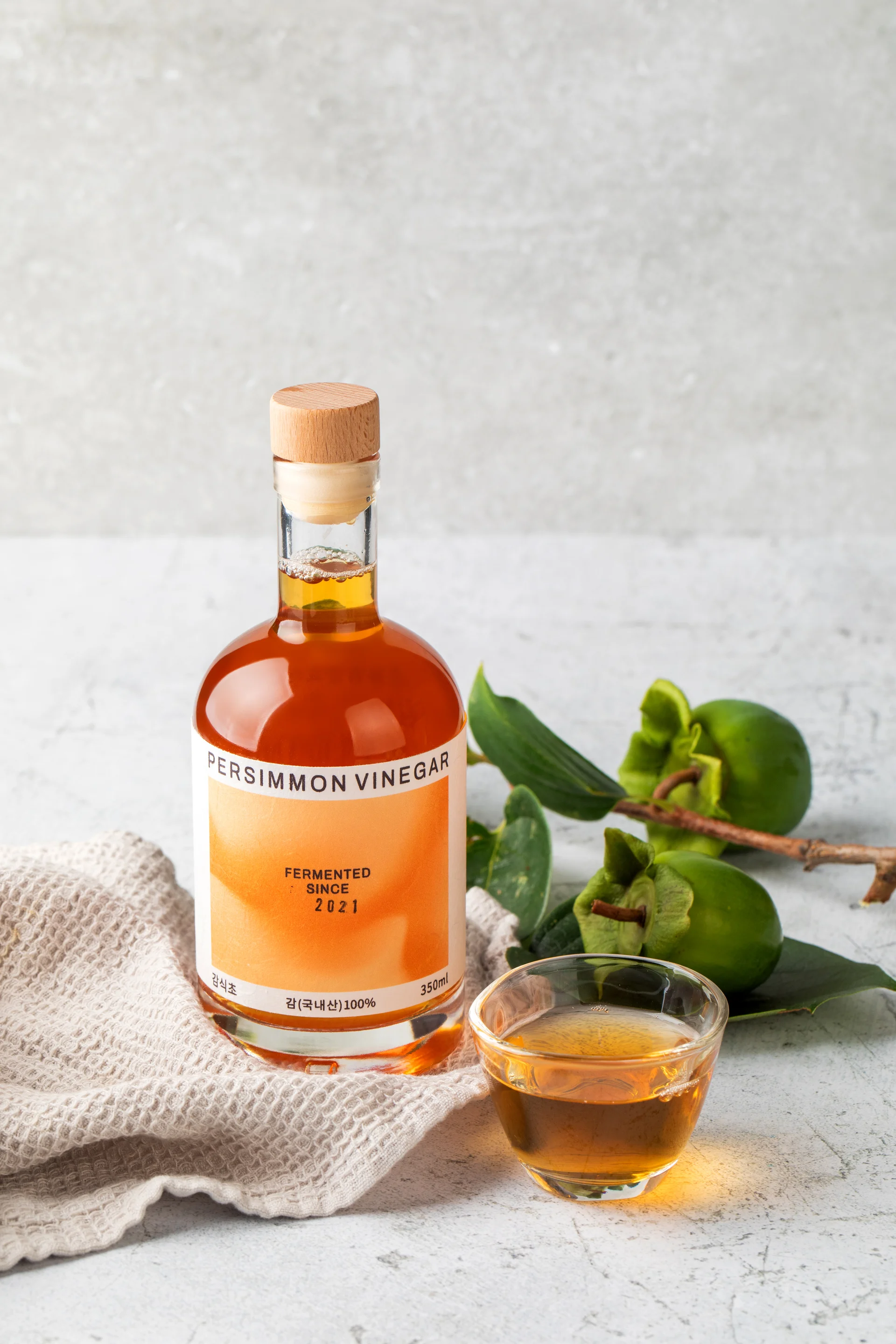 은가비 브랜드의 발효식초 3종(사과식초, 감식초, 식초 음료)이 신선한 사과와 감 원물과 함께 연출된 고감도 촬영 이미지. 한여름 감 수급 변수를 극복하고 기획자가 직접 디렉팅한 내추럴 톤의 브랜드 비주얼 컷.
