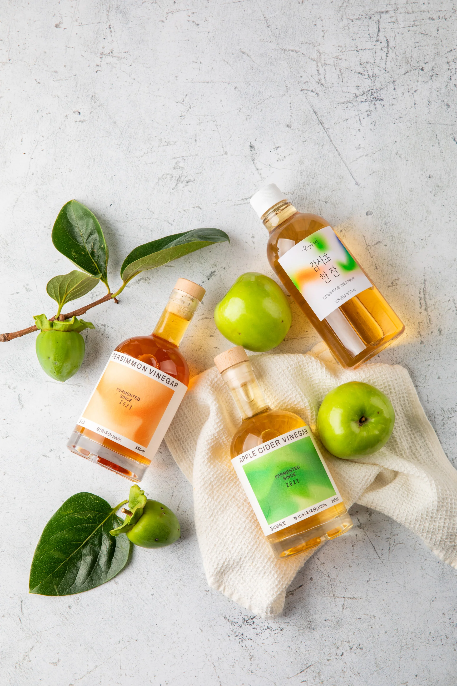 은가비 브랜드의 발효식초 3종(사과식초, 감식초, 식초 음료)이 신선한 사과와 감 원물과 함께 연출된 고감도 촬영 이미지. 한여름 감 수급 변수를 극복하고 기획자가 직접 디렉팅한 내추럴 톤의 브랜드 비주얼 컷.
