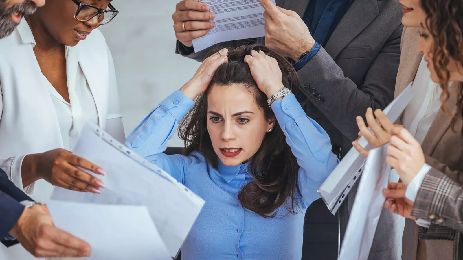 사무실에서 압도된 여성을 보여주는 하이앵글 사진. 파란색 셔츠를 입은 한 여성이 머리를 쥐어뜯으며 괴로워하고 있고, 그녀 주위의 5명의 동료들이 그녀에게 종이 서류 뭉치를 밀어붙이며 압박하고 있다.