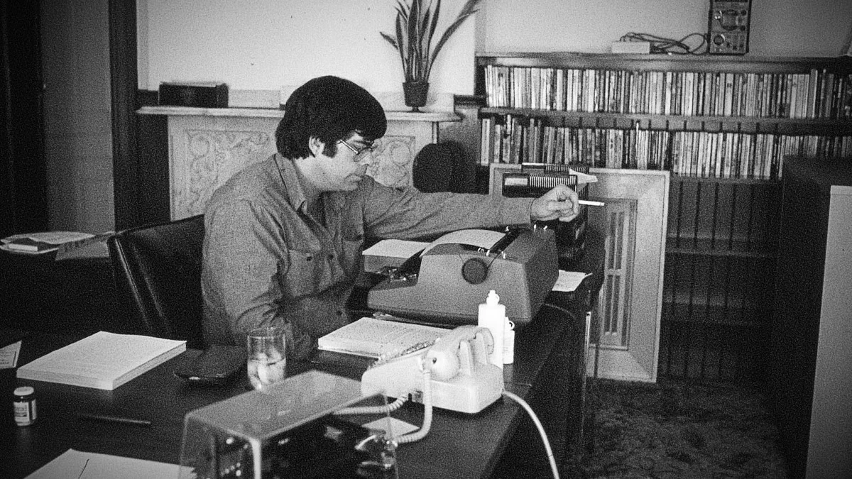 Black-and-white photograph of Stephen King writing at a desk with a typewriter, surrounded by books and papers in a quiet room.