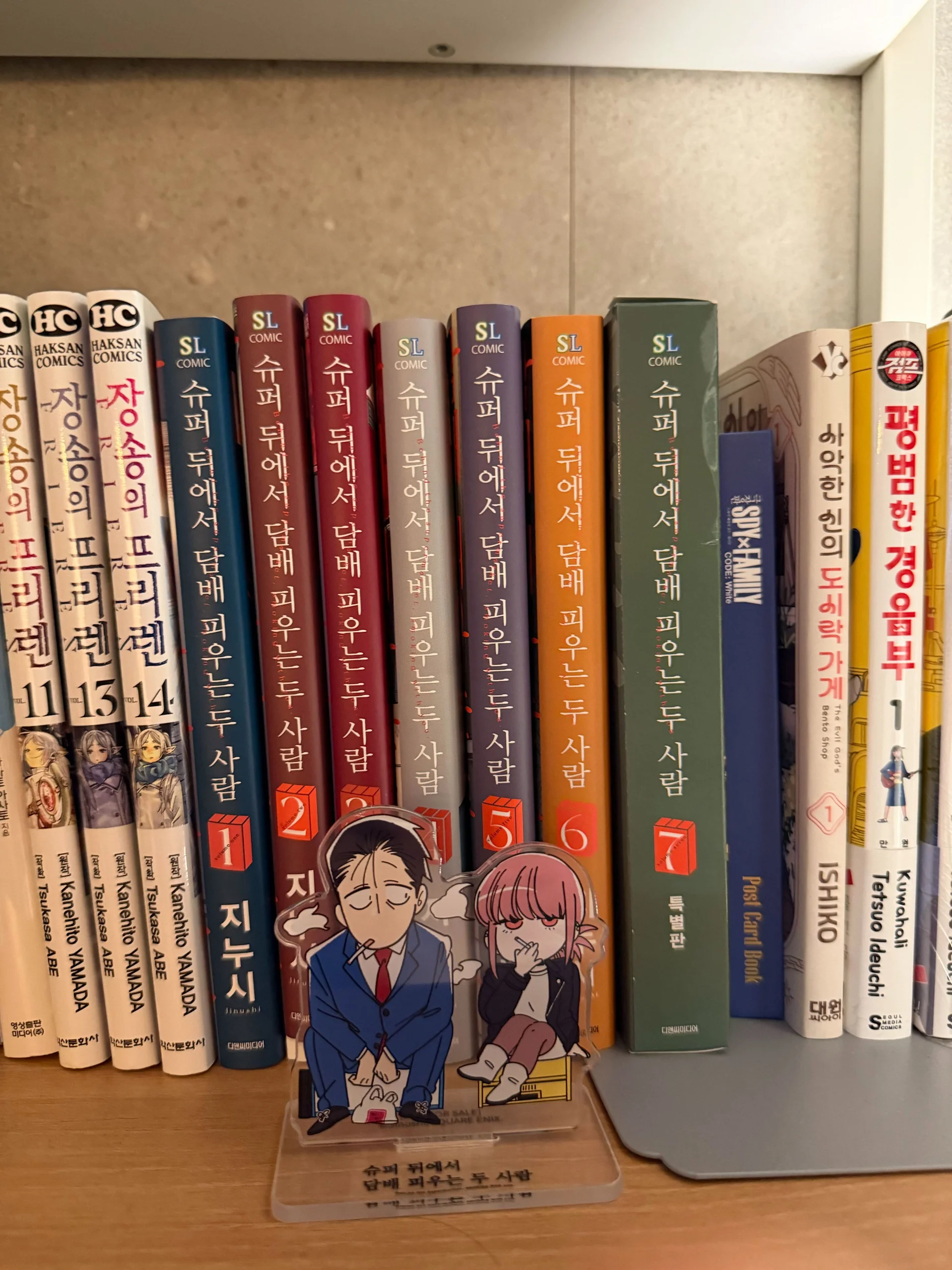 책장 위에 일본 만화와 한국어 번역 만화책이 꽂혀 있는 사진. 가운데에는 만화 『슈퍼 뒤에서 담배 피우는 두 사람』 1권부터 7권까지 여러 권이 나란히 꽂혀 있고, 앞쪽에는 남성과 여성 캐릭터가 함께 앉아 담배를 피우는 아크릴 스탠드 굿즈가 놓여 있다. 왼쪽에는 『장송의 프리렌』 만화책 여러 권이 보이며, 오른쪽에는 다른 일본 만화 단행본들이 함께 진열되어 있다. 만화책 수집, 일본 만화 추천, 『슈퍼 뒤에서 담배 피우는 두 사람』 굿즈와 책장 디스플레이를 보여주는 사진.