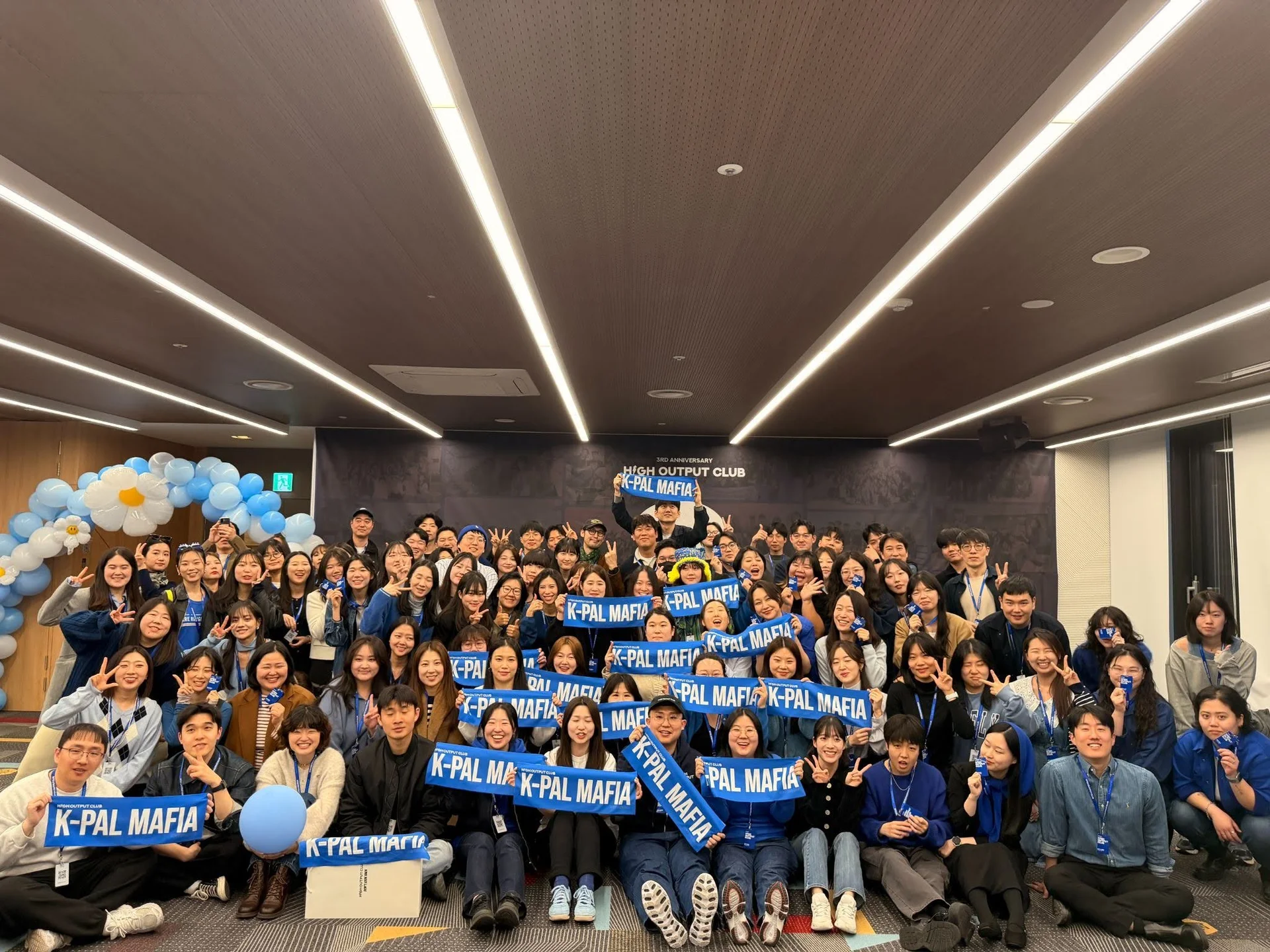 하이아웃풋클럽 3주년 행사에서 멤버들이 파란색 'K-PAL MAFIA' 배너를 들고 단체사진을 찍고 있다. 사원증 목걸이를 착용한 멤버들이 보인다.