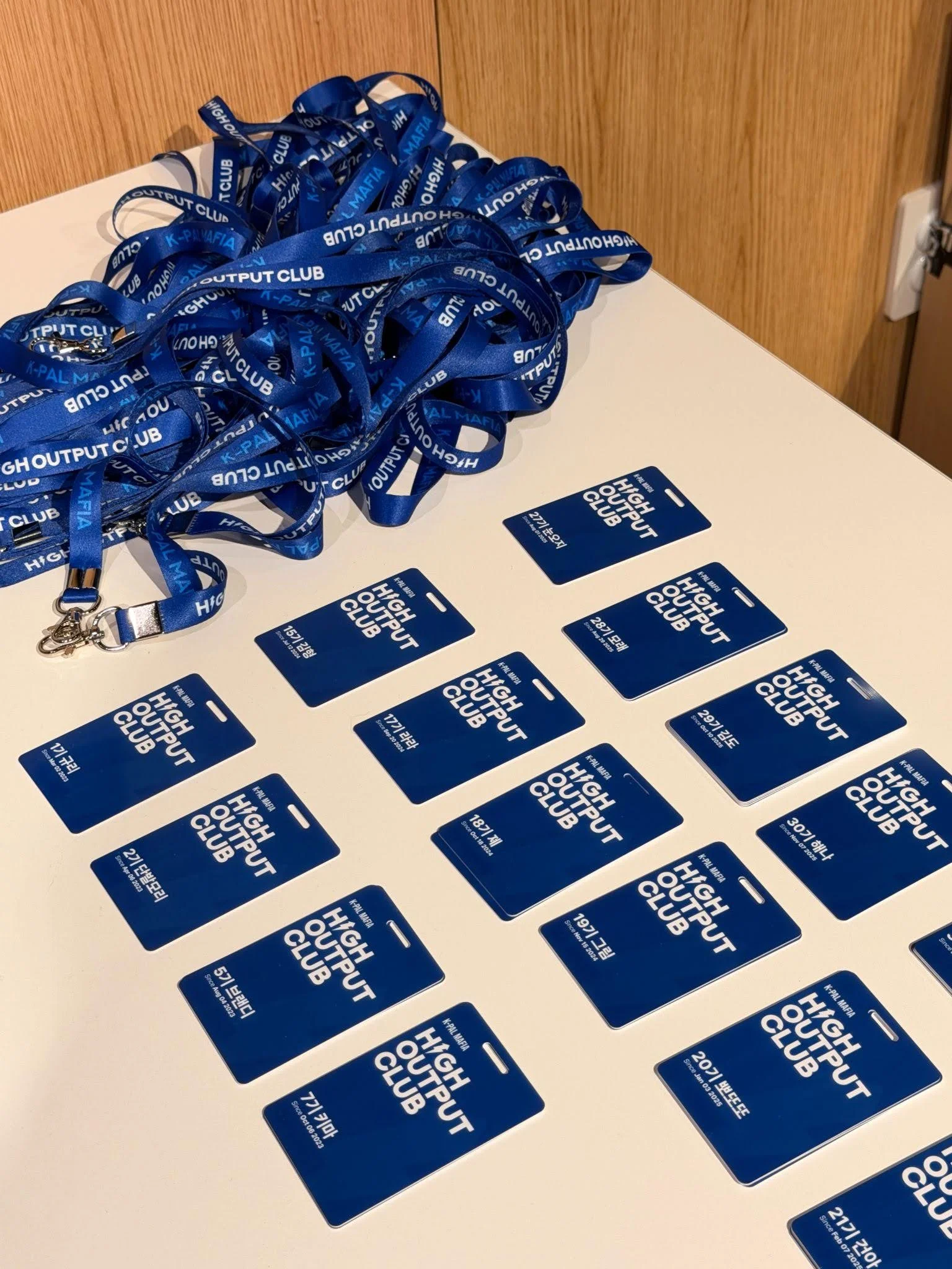 테이블 위에 파란색 HOC 사원증이 기수와 이름별로 나열되어 있다. 'K-PAL MAFIA HIGH OUTPUT CLUB' 로고가 적힌 카드와 파란색 넥스트랩이 함께 놓여 있다.
