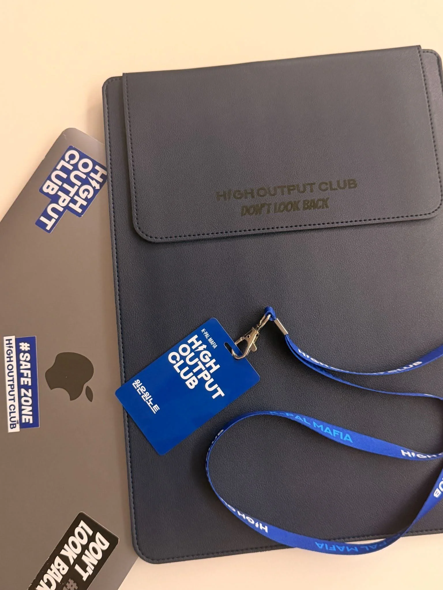  'HIGH OUTPUT CLUB DON'T LOOK BACK' 문구가 새겨진 네이비 노트북 파우치 위에 파란색 사원증과 넥스트랩이 놓여 있고, 옆에 HOC 스티커가 함께 있다.