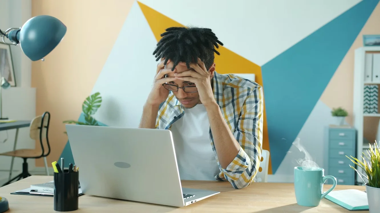 A man is sitting at a desk in front of a laptop. He looks tired or stressed. 