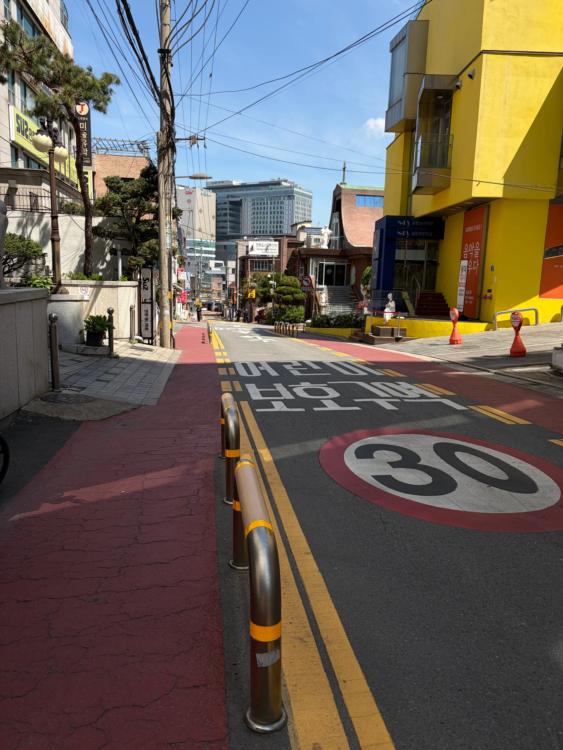 A sunny sloped street in Korea, with road markings and colorful buildings on both sides.