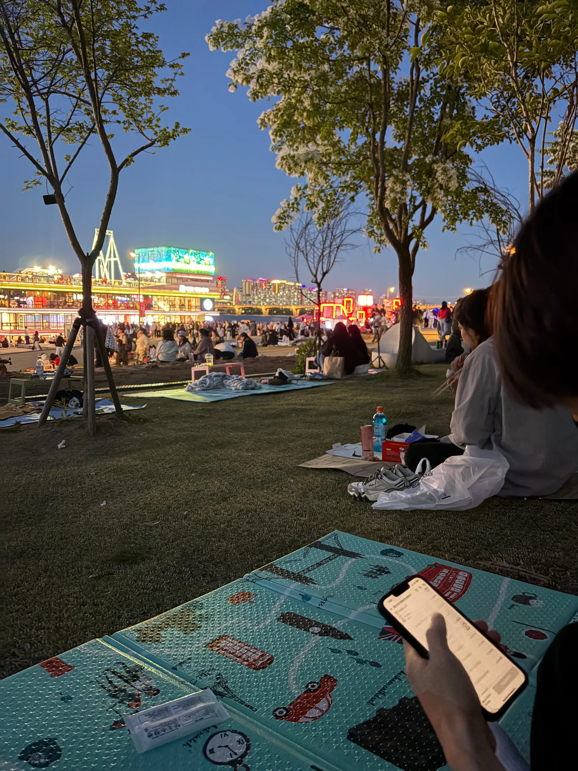 A lively evening scene by the river in Korea, with people sitting on the grass, city lights glowing in the background, and a relaxed, social atmosphere.
