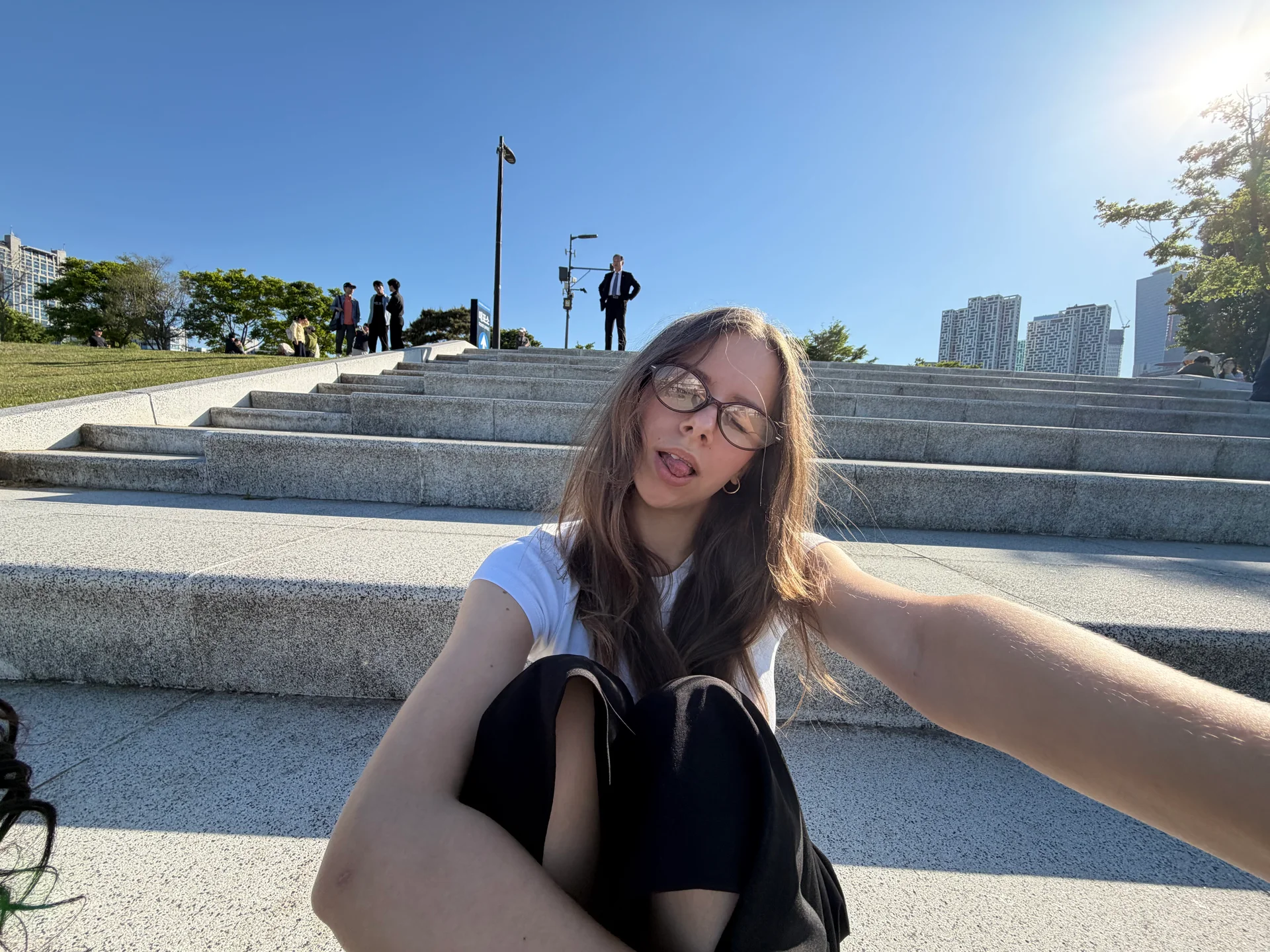 A young woman sitting on wide outdoor steps under a bright blue sky, taking a relaxed selfie while people stand and walk in the background.