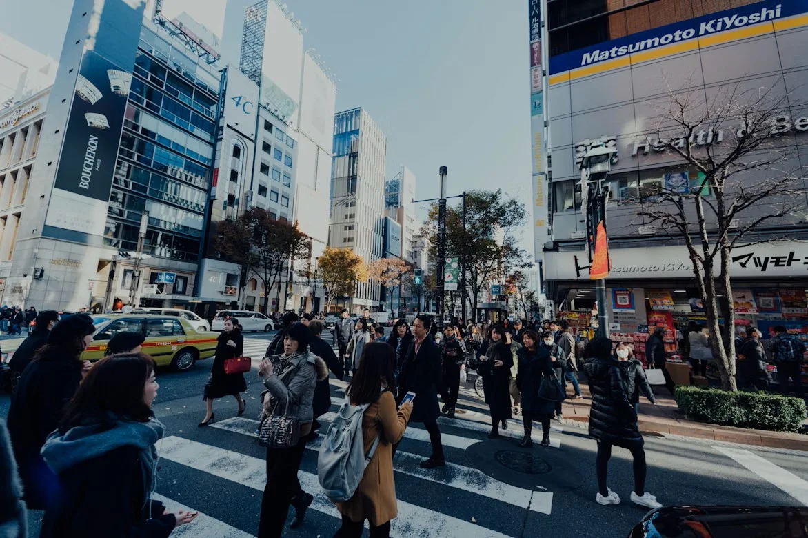 A busy street in Japan with many people crossing the road between tall buildings on a bright day.
