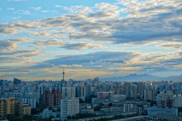 A large city skyline under a cloudy blue sky, with many buildings and mountains in the distance.