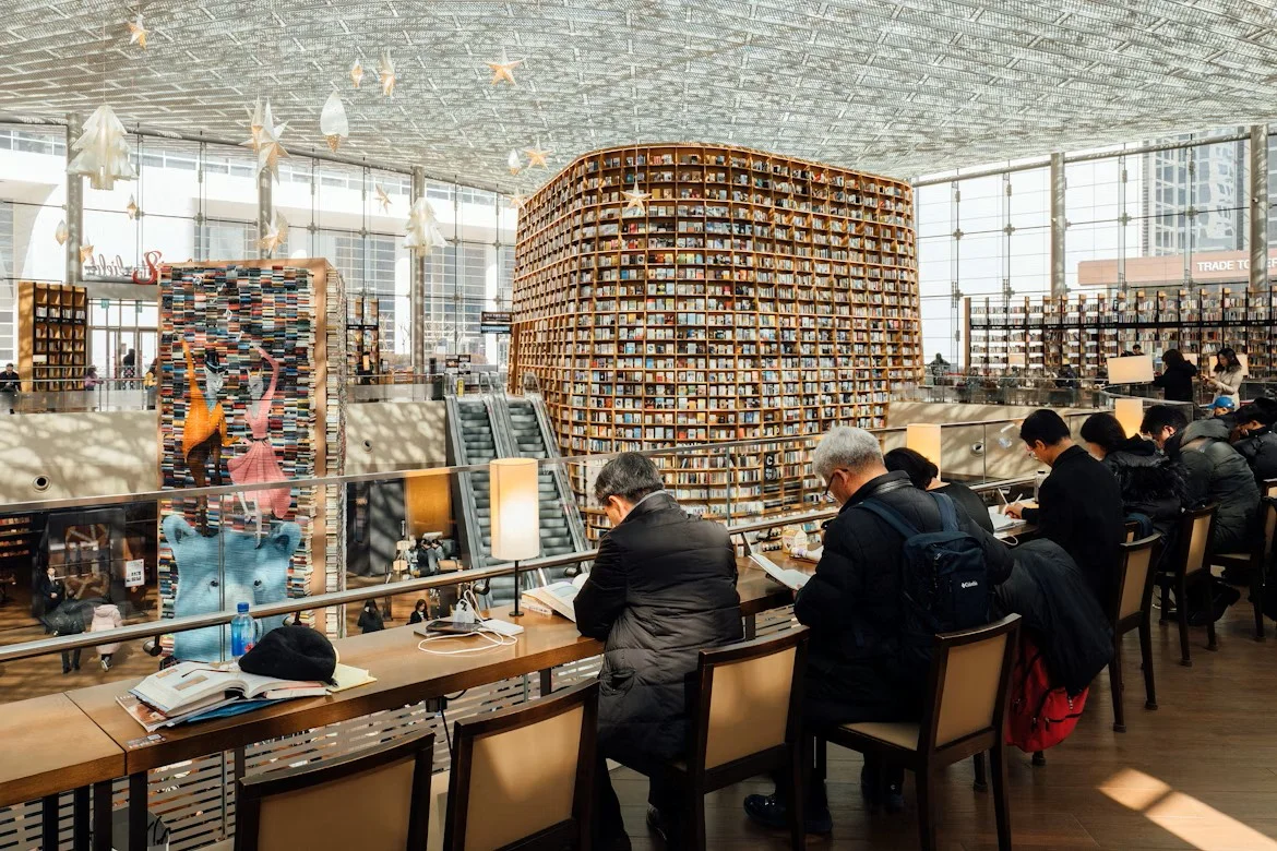 A large, modern library in Seoul, filled with towering bookshelves and people reading or working in a calm, bright atmosphere.