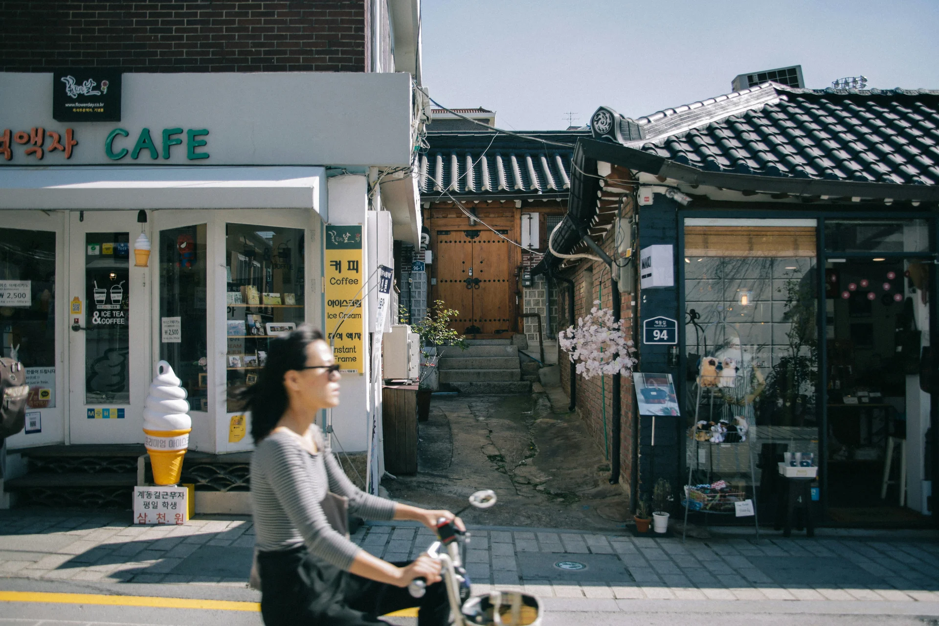 A woman rides past small cafés and traditional buildings on a quiet street in Seoul.