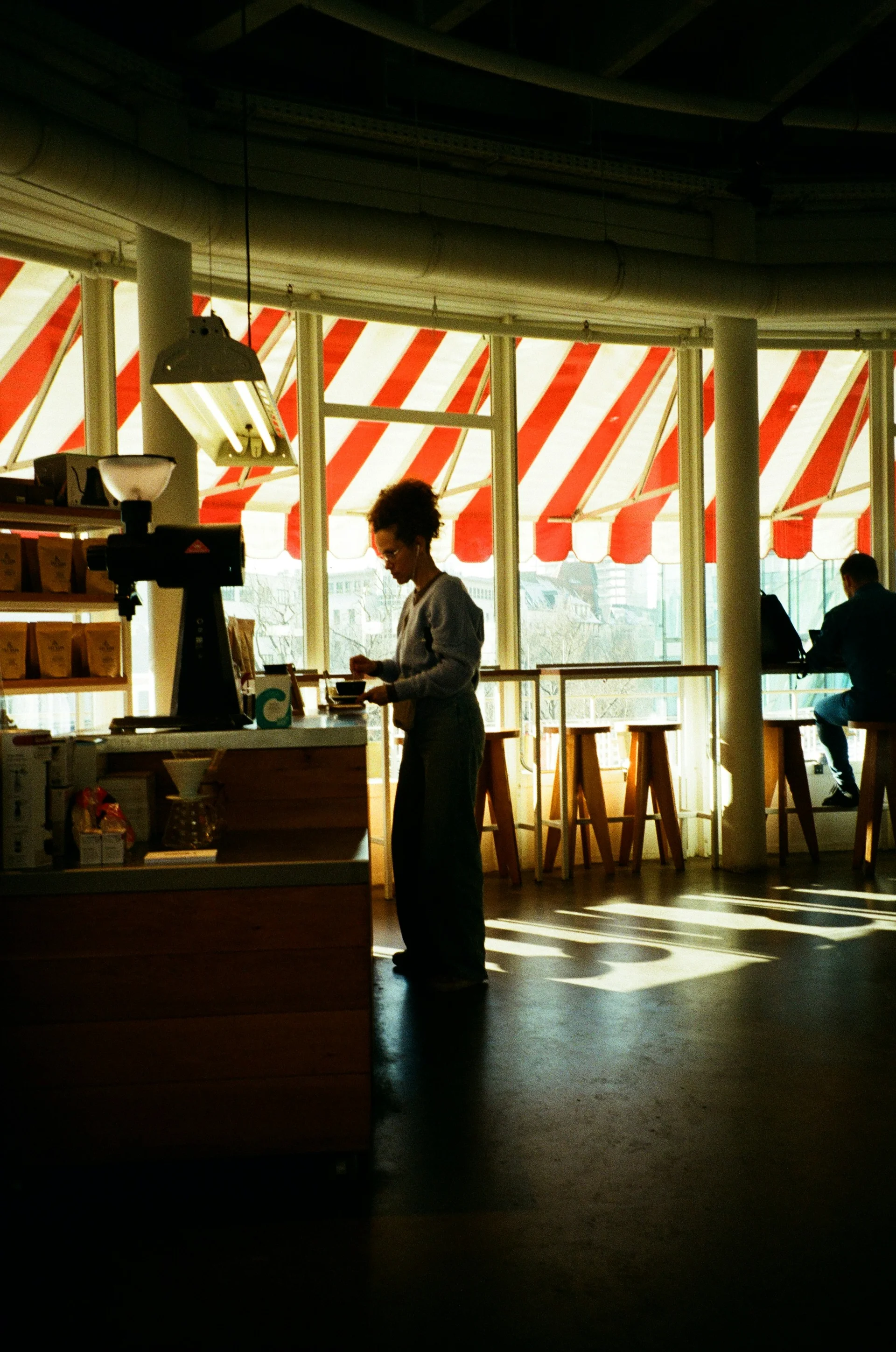 A woman stands in a sunlit café while people sit quietly by the windows.