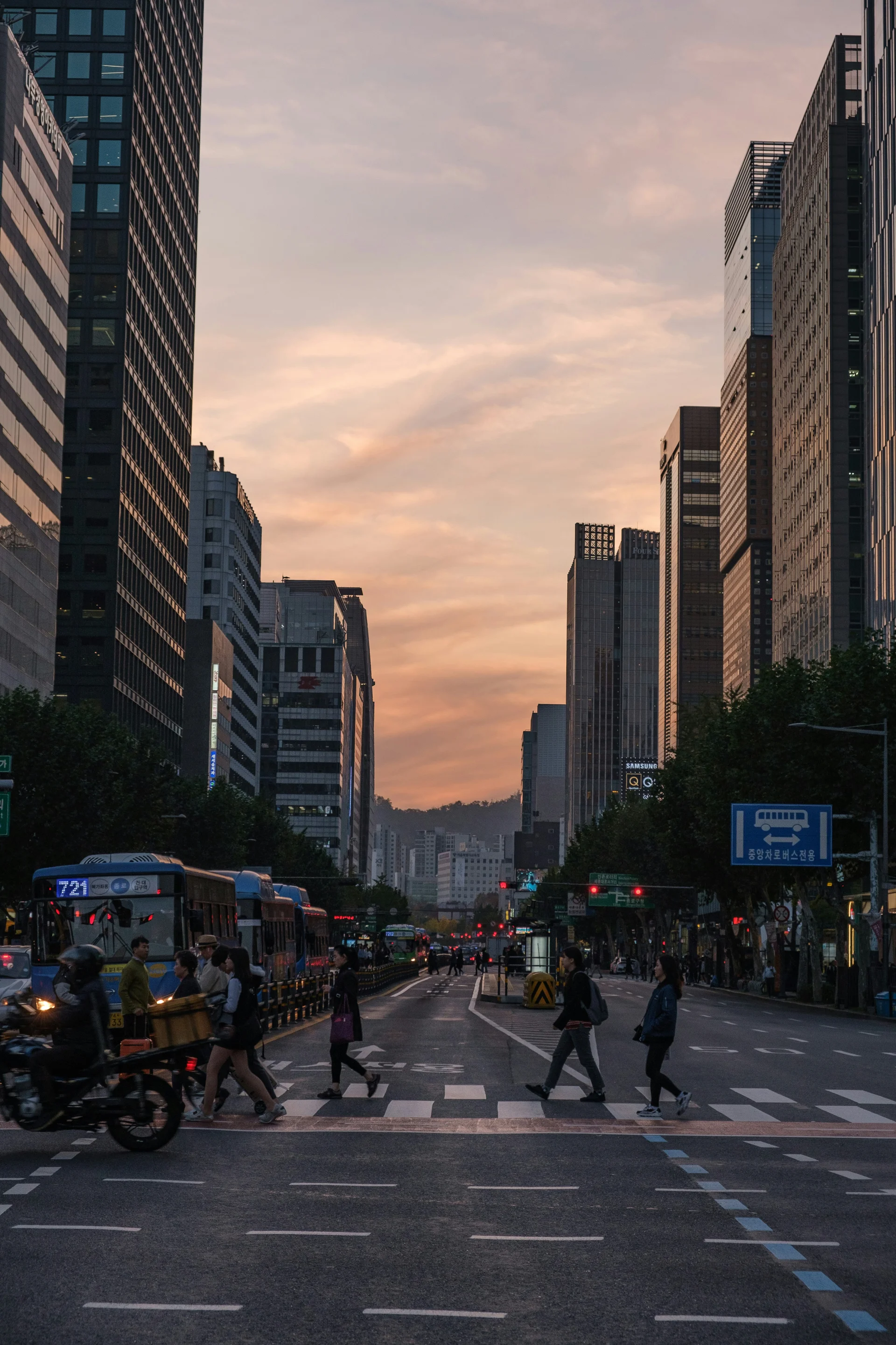 People cross a wide city street between tall buildings at sunset.