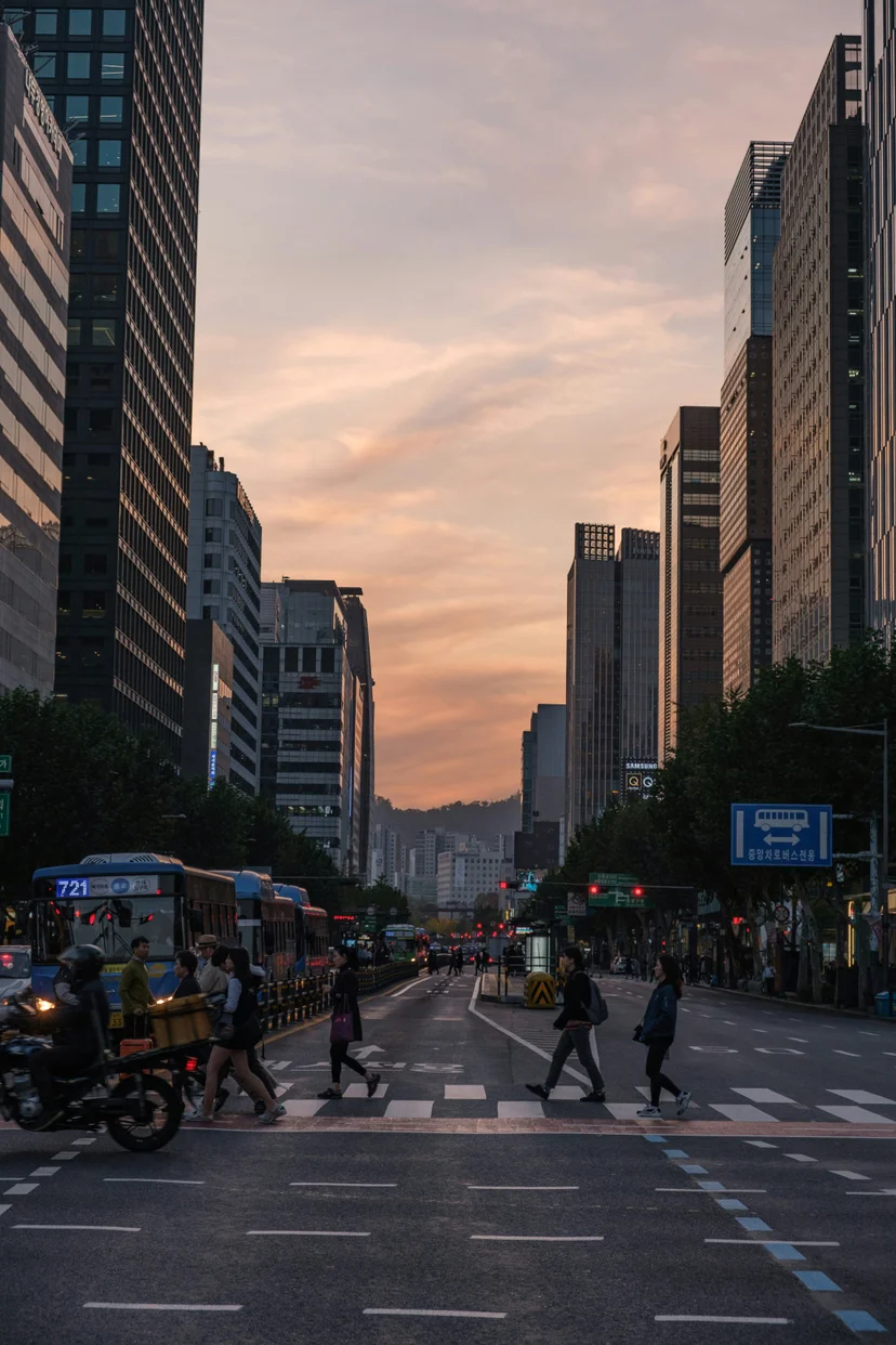 People cross a wide city street between tall buildings at sunset.