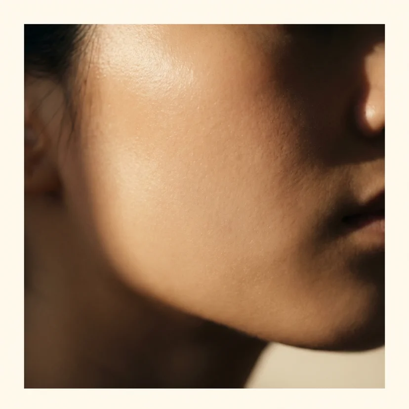 Close-up of a woman's cheek and jawline showing smooth, even-textured skin with a natural dewy finish, photographed in warm directional light against a soft cream background