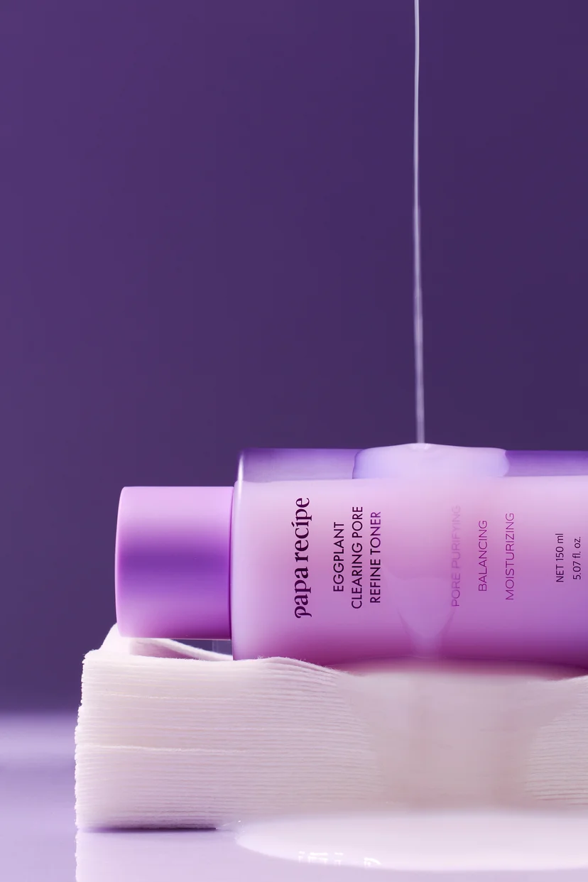 Papa Recipe Eggplant Clearing Pore Refine Toner 150ml bottle lying on its side on a stack of cotton pads, with a thin stream of milky toner pouring down onto the bottle from above, against a deep purple background